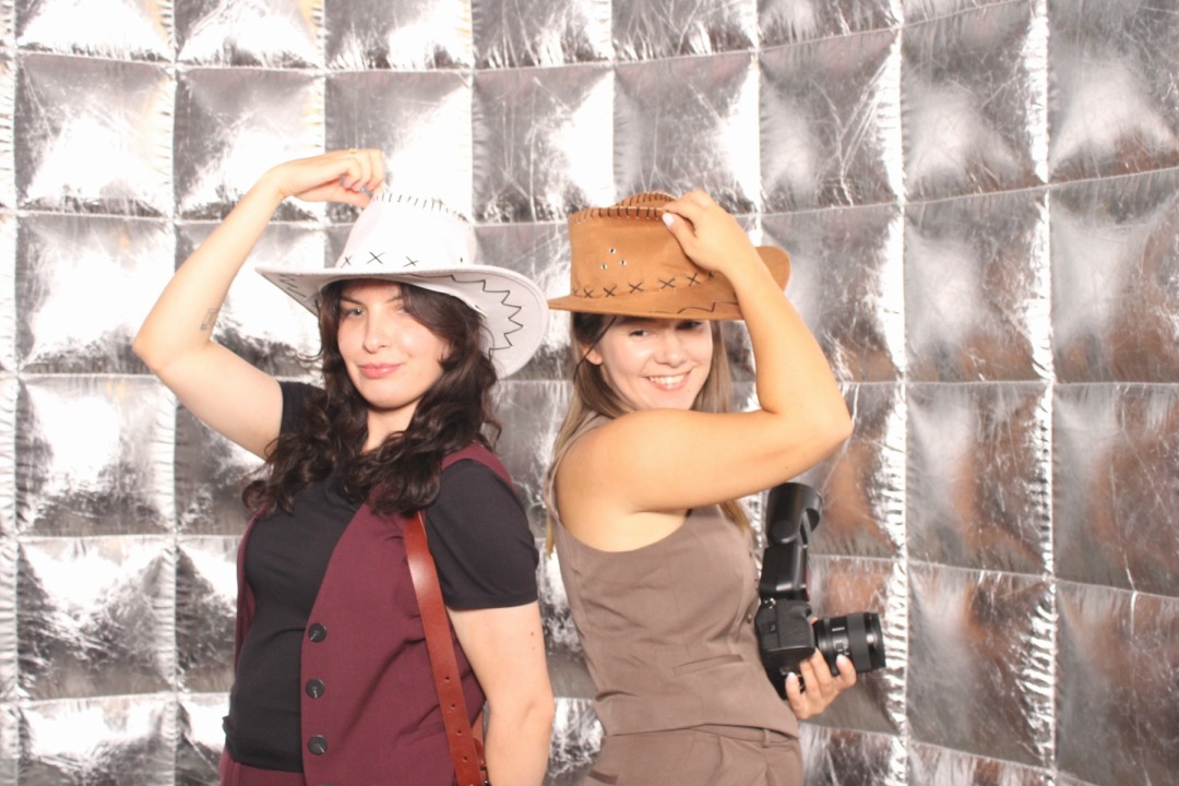 Dwie uśmiechnięte kobiety w kowbojskich kapeluszach pozują na tle srebrnej ścianki w Porębie Wielkiej. Jedna trzyma aparat z lampą błyskową.