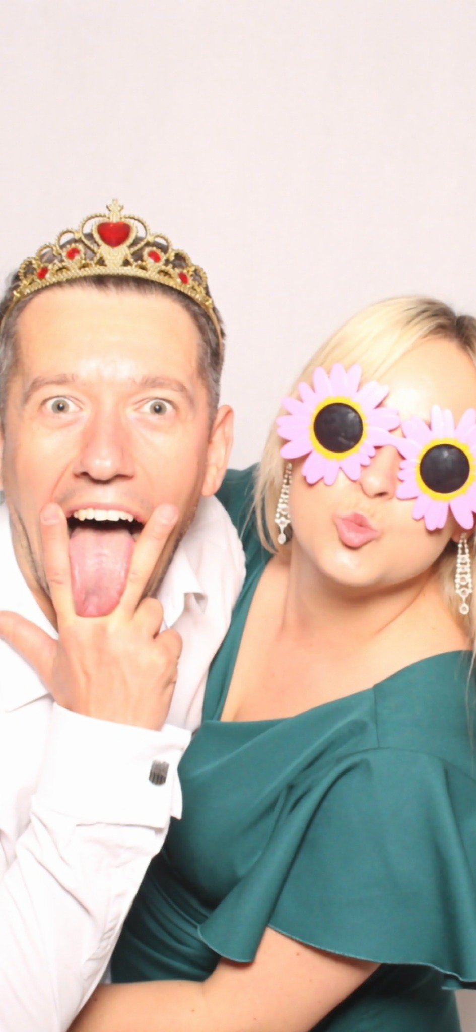 Para w fotobudce: mężczyzna w koronie pokazuje język, kobieta w okularach w kształcie kwiatów robi dzióbek. Białe tło, radosna atmosfera.