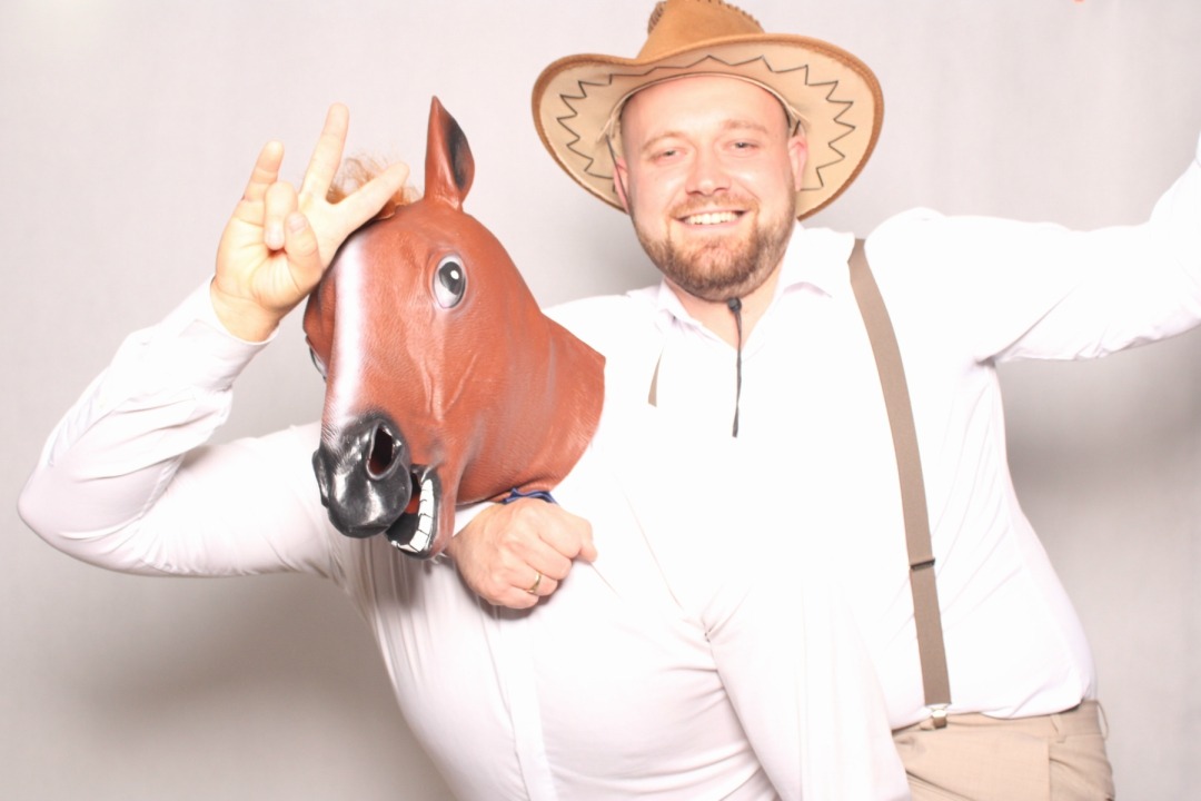 Uśmiechnięty mężczyzna w kowbojskim kapeluszu pozuje z osobą w masce konia, pokazując znak pokoju. Zabawna fotografia z imprezy, jasne tło.