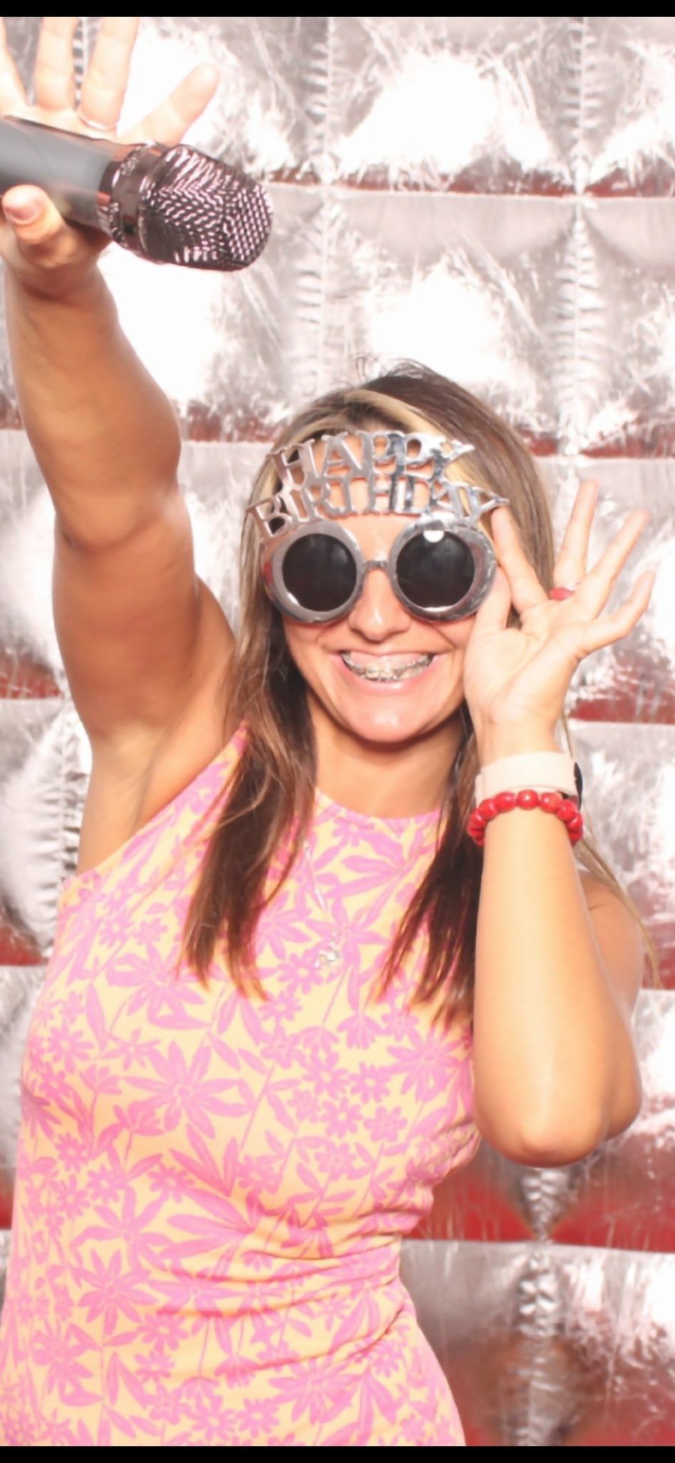 Uśmiechnięta kobieta w okularach 'Happy Birthday' i kwiecistej sukience pozuje z mikrofonem na srebrnym tle, prawdopodobnie w fotobudce.