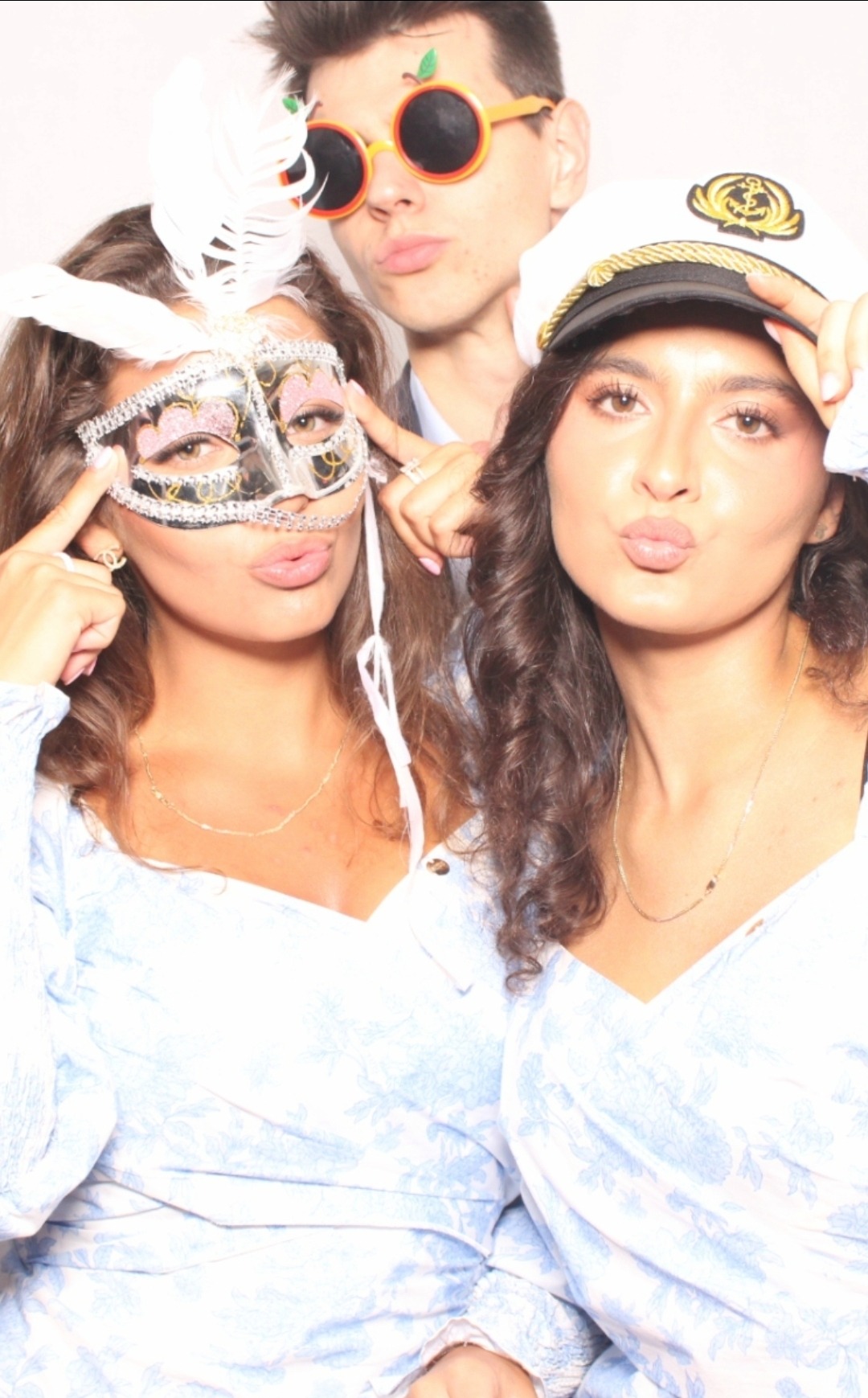 Trzy osoby pozują do zdjęcia w fotobudce, ubrane w zabawne rekwizyty: maskę karnawałową, kapitańską czapkę i pomarańczowe okulary z jabłkami.