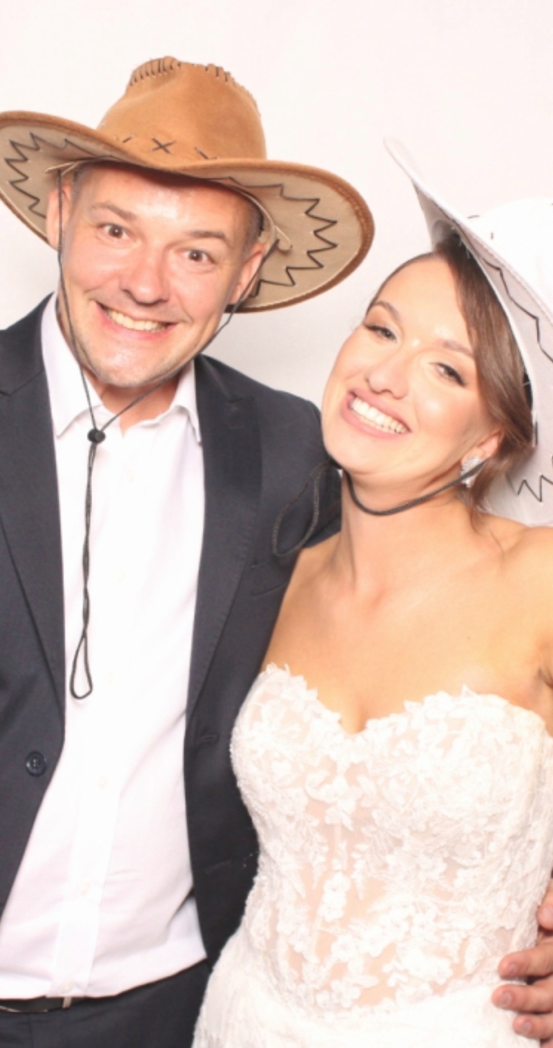 Uśmiechnięta para w strojach kowbojskich pozuje do zdjęcia na tle białej ścianki. Pan w garniturze, pani w koronkowej sukni ślubnej i kapeluszu.