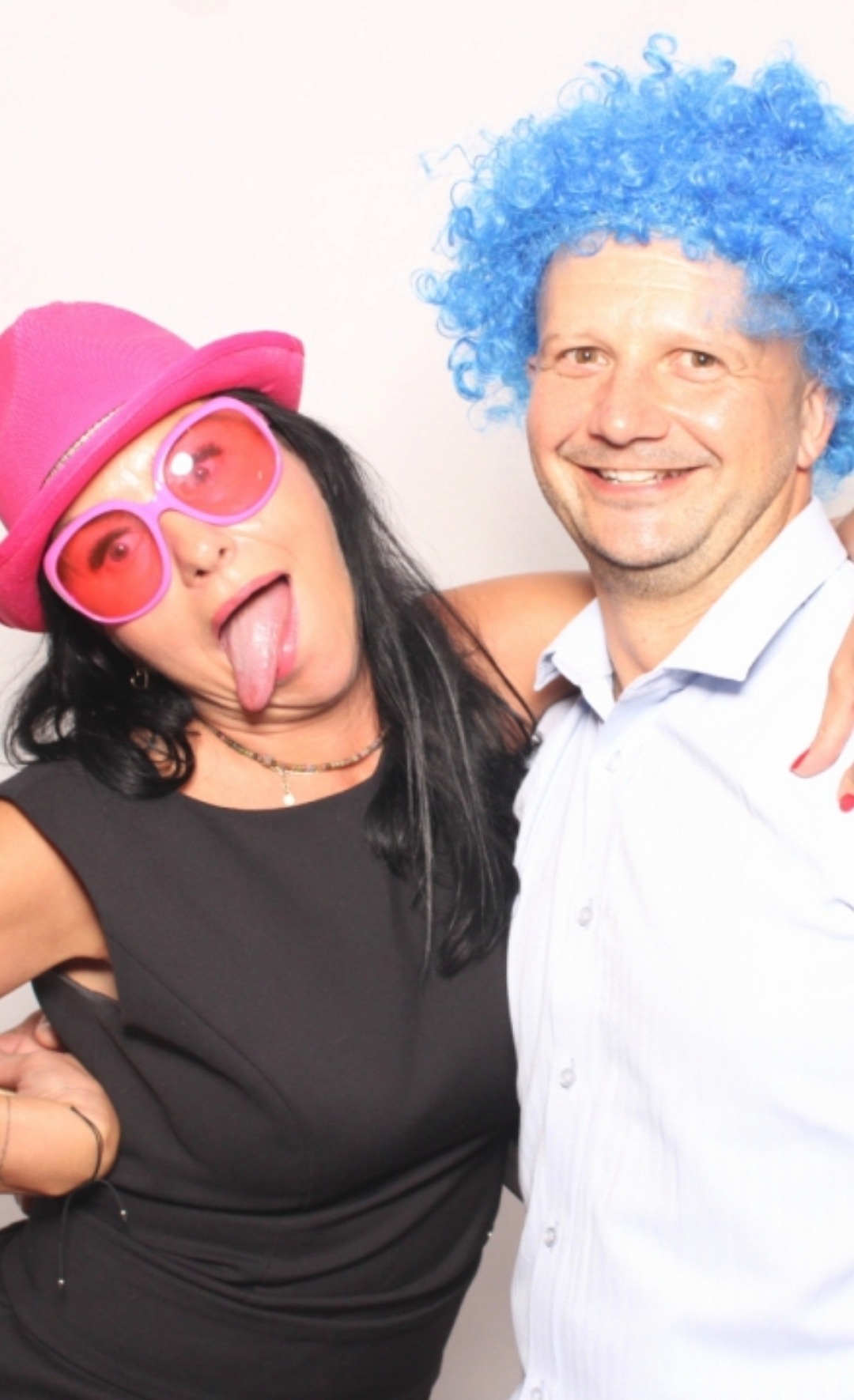 Dwie osoby w przebraniach: kobieta w różowym kapeluszu i okularach wystawia język, mężczyzna w niebieskiej peruce uśmiecha się do kamery. Zabawa w fotobudce.