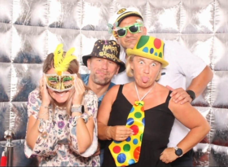 Grupa czterech osób w przebraniach pozuje w fotobudce z srebrnym tłem. Uśmiechy, maski, kapelusze i krawat w kropki dodają wesołego charakteru.