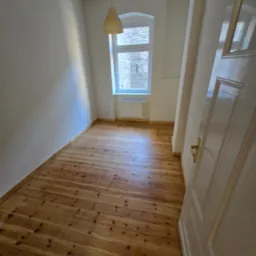 Jasne, odnowione deski podłogowe w pokoju z oknem łukowym i wiszącą lampą. Częściowo otwarte białe drzwi z złotą klamką po prawej stronie.