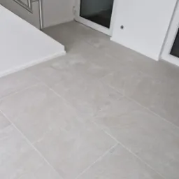 Jasnoszare płytki podłogowe ułożone w korytarzu z metalową balustradą schodów i białymi drzwiami wejściowymi. Minimalistyczny design, czyste linie i neutralna kolorystyka.