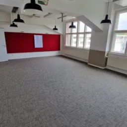 Przestronne biuro z szarą wykładziną dywanową, czerwonym panelem ściennym i wiszącymi lampami. Widok na okna z grzejnikami. Białe ściany i sufit z instalacjami.