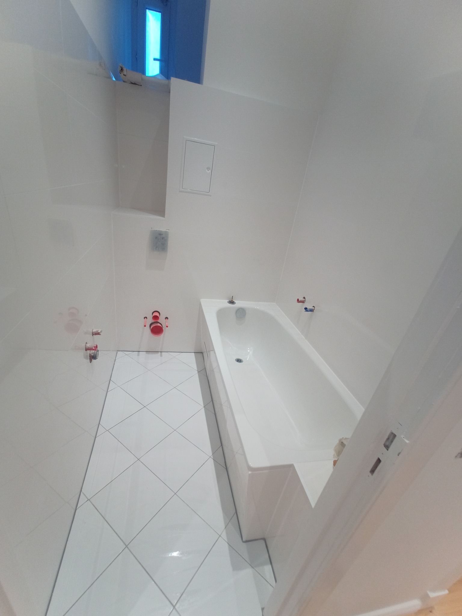 Biała łazienka po remoncie: wanna, płytki podłogowe ułożone w karo, świeżo wykończone ściany z instalacją sanitarną. Minimalistyczny design z niebieskim akcentem okna.