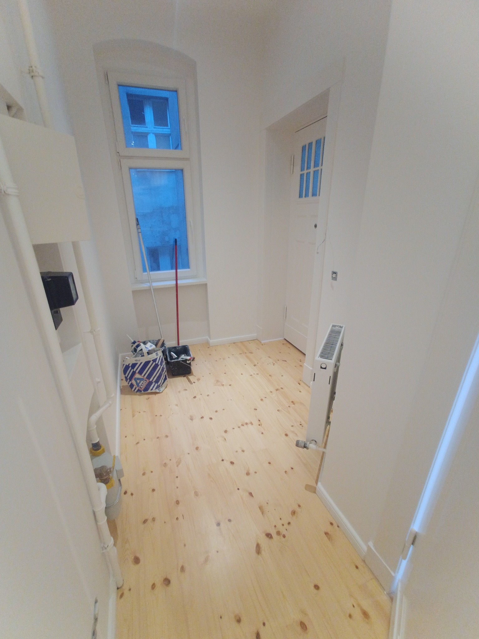 Jasne, odnowione drewniane podłogi w korytarzu z białymi ścianami i drzwiami. Widoczne akcesoria malarskie przy oknie. Ujęcie z perspektywy podłogi.