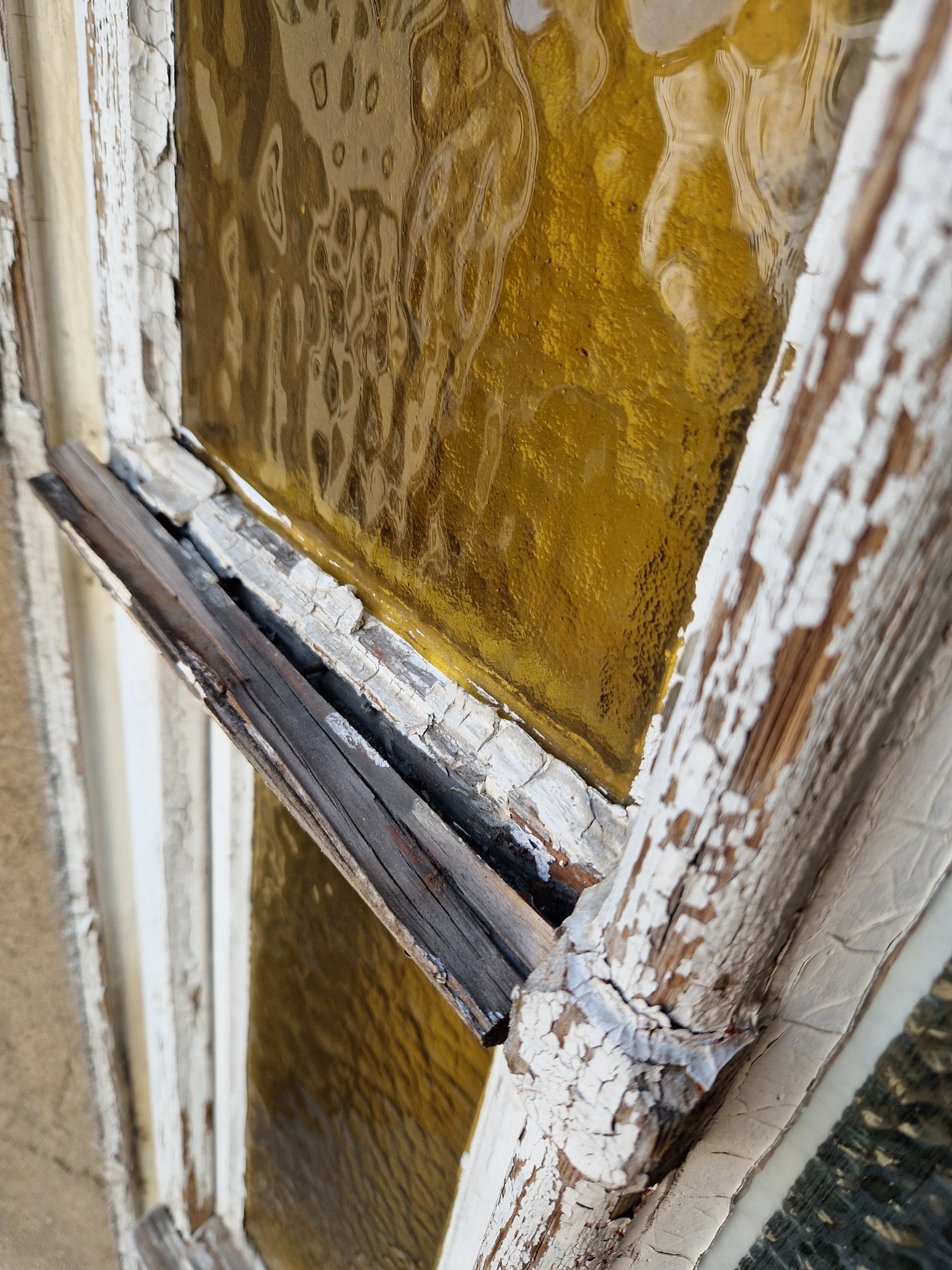Zniszczone, stare okno z żółtą szybą i łuszczącą się białą farbą. Widoczne uszkodzenia ramy i detale faktury drewna. Perspektywa z dołu, ukazująca zły stan techniczy.