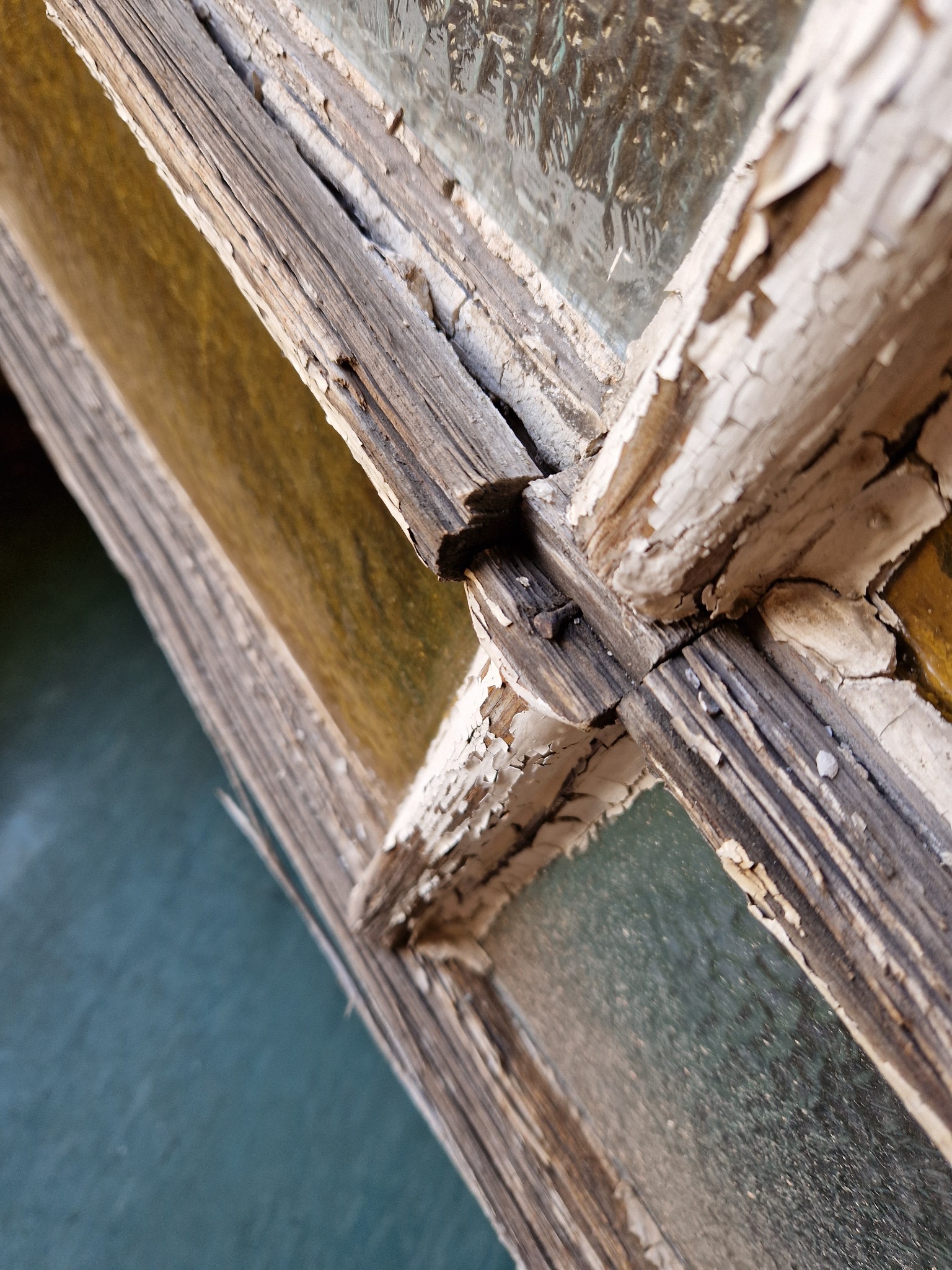 Fragment starego, drewnianego okna z łuszczącą się białą farbą, widoczne słoje drewna i metalowy gwóźdź. Perspektywa z dołu.