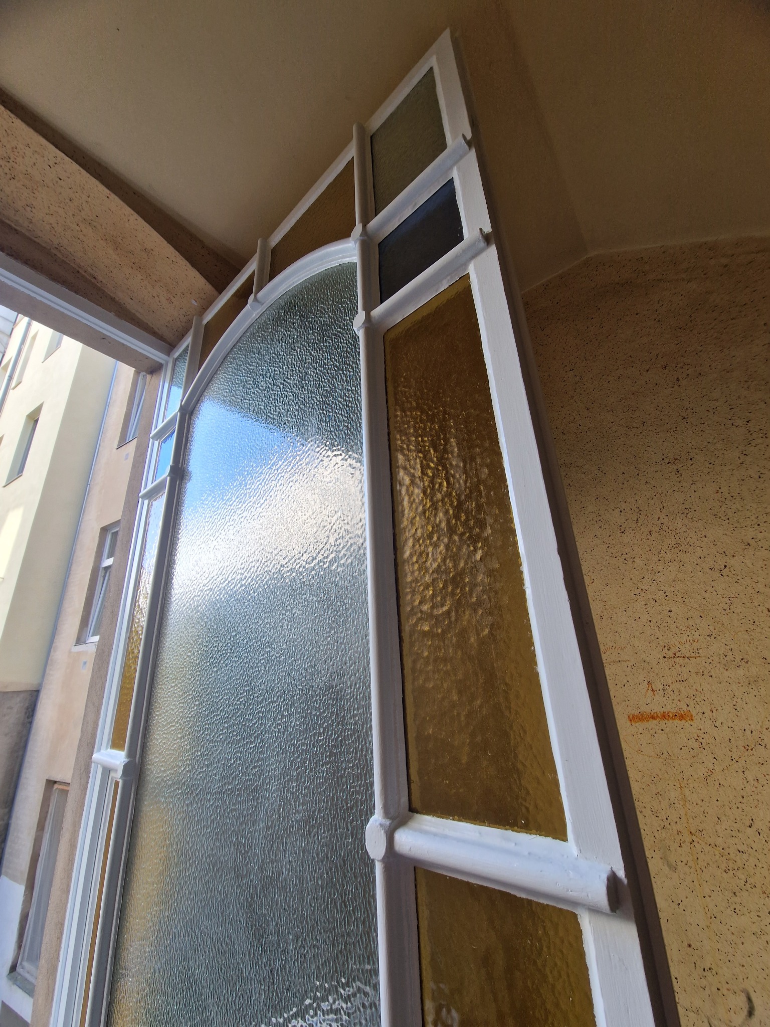 Szklane, zdobione drzwi z białymi ramami, widziane od wewnątrz, z widokiem na budynek na zewnątrz. Drzwi z kolorowymi szybkami i wzorzystym szkłem.