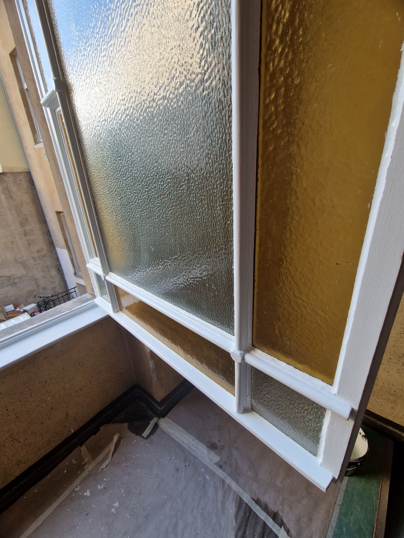 Otwarty, odnowiony fragment okna z ozdobnym szkłem w białej ramie, widok z góry, z fragmentem parapetu i śladami prac malarskich na podłodze.