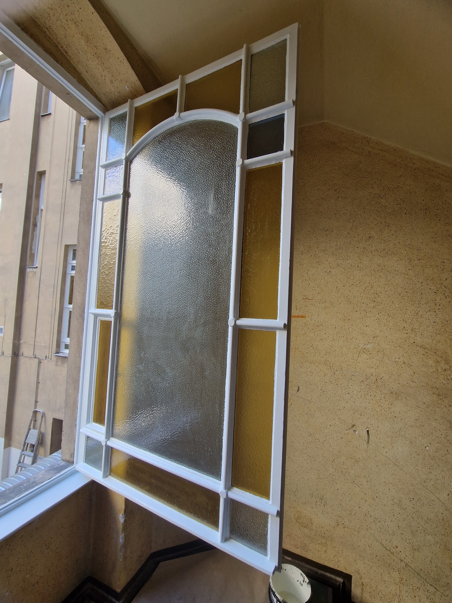 Odnowione okno witrażowe w kamienicy w Świebodzinie. Biała rama, ozdobne szkło i żółte akcenty. Widok z balkonu, ściana o fakturze baranka.