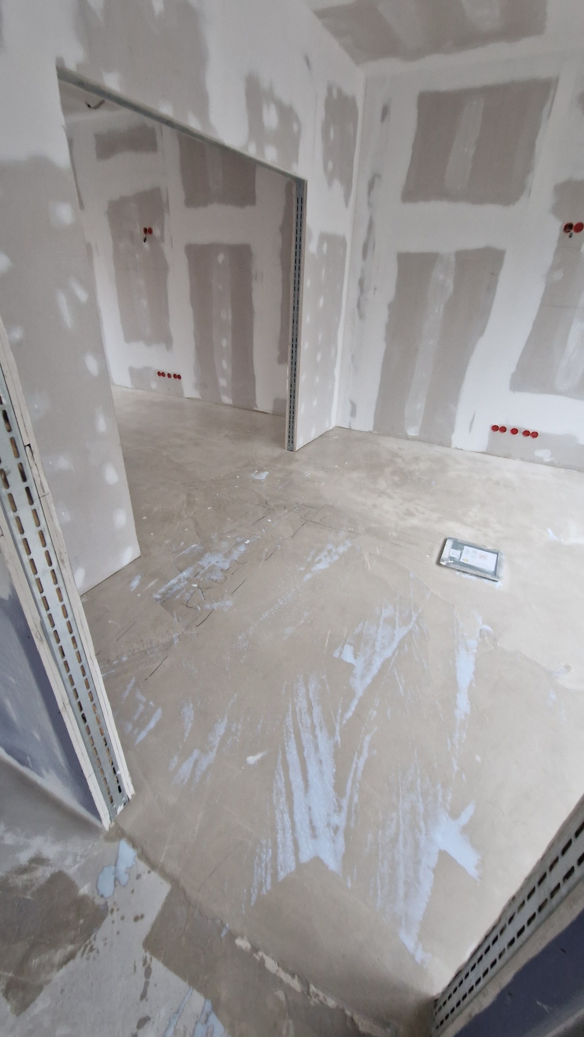 Surowe wnętrze w trakcie prac wykończeniowych: ściany z płyt gipsowo-kartonowych, wylewka podłogowa z widocznymi śladami szpachli, metalowe profile ościeżnic.