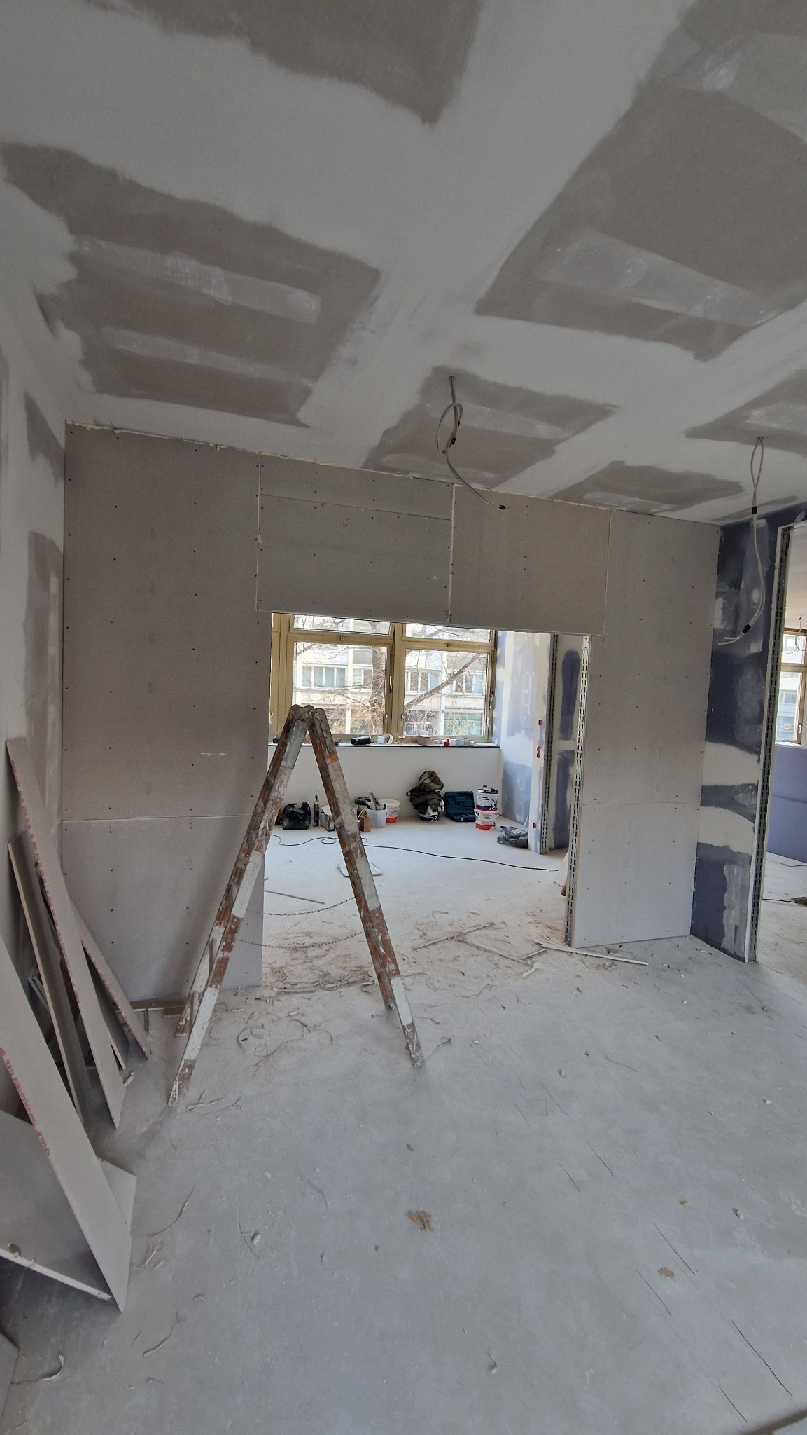 Pokój w trakcie prac. Widoczne płyty gipsowo-kartonowe na ścianach i suficie, drabina malarska i narzędzia. Okno w tle. Etap szpachlowania sufitu.