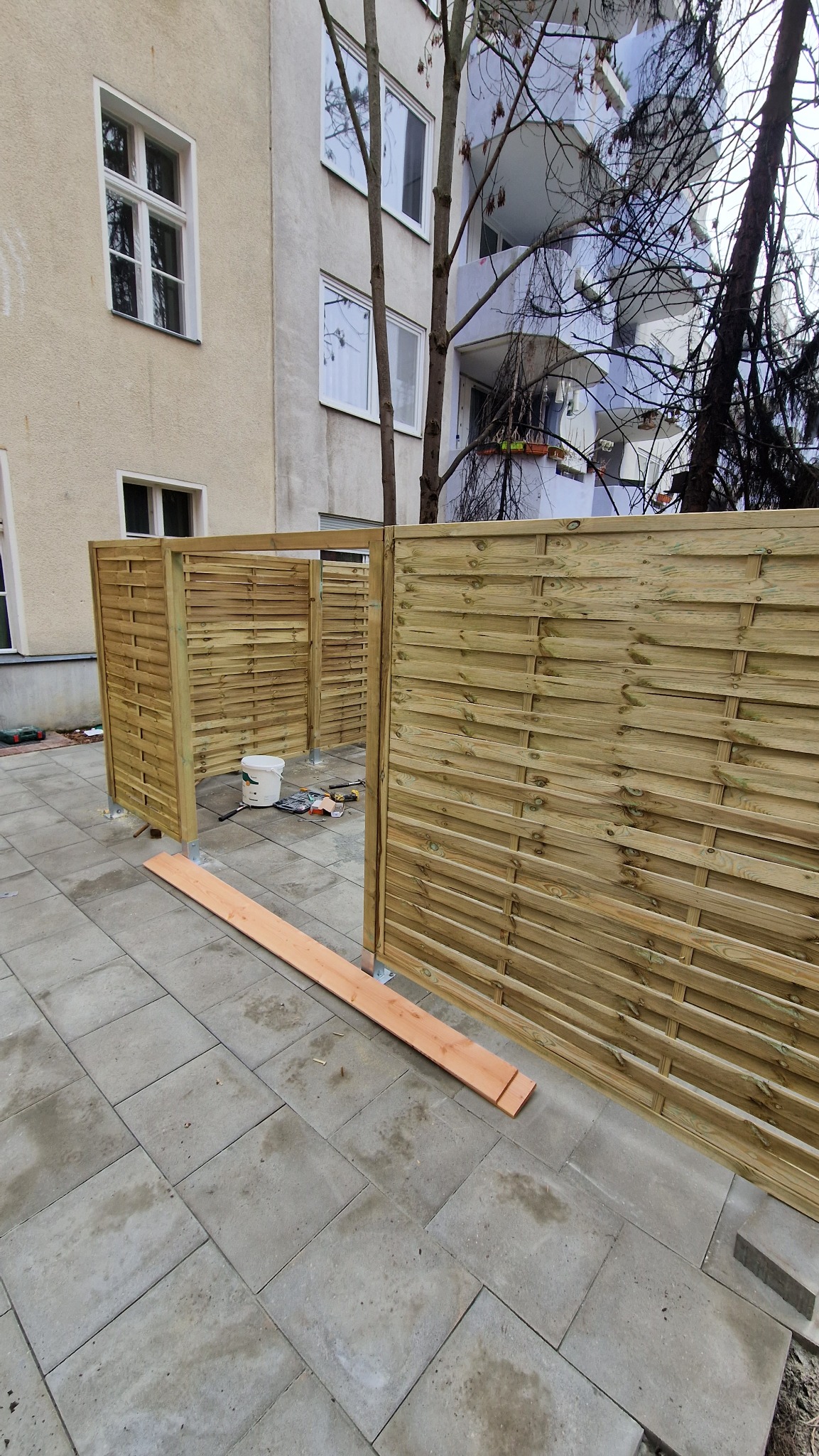 Montaż ażurowego, drewnianego ogrodzenia panelowego na betonowej kostce brukowej przy bloku. Widoczne narzędzia i wiadro farby.