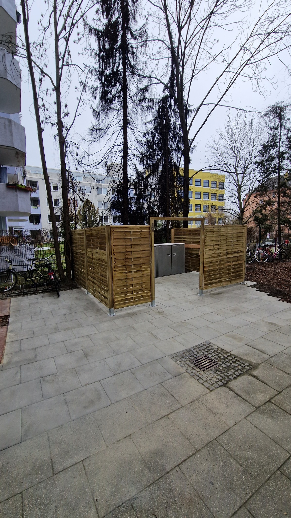 Nowoczesna zabudowa śmietnikowa z drewnianym ogrodzeniem panelowym na betonowej nawierzchni, z widocznymi drzewami i budynkami w tle.