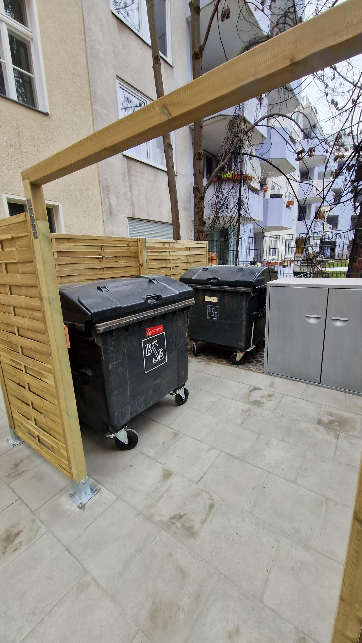 Drewniana osłona śmietników z panelami i metalowymi mocowaniami na jasnej kostce brukowej, z widocznymi pojemnikami na odpady i budynkami w tle.