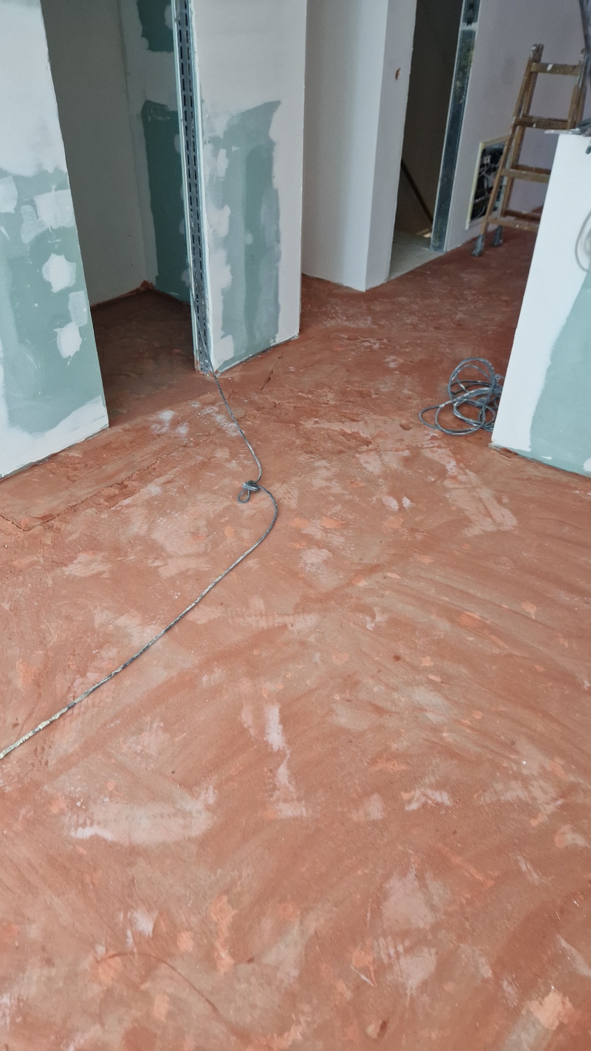 Surowy stan wnętrza w trakcie prac wykończeniowych: częściowo zagruntowana podłoga, ściany z płyt gipsowo-kartonowych, metalowe profile, drabina w tle.