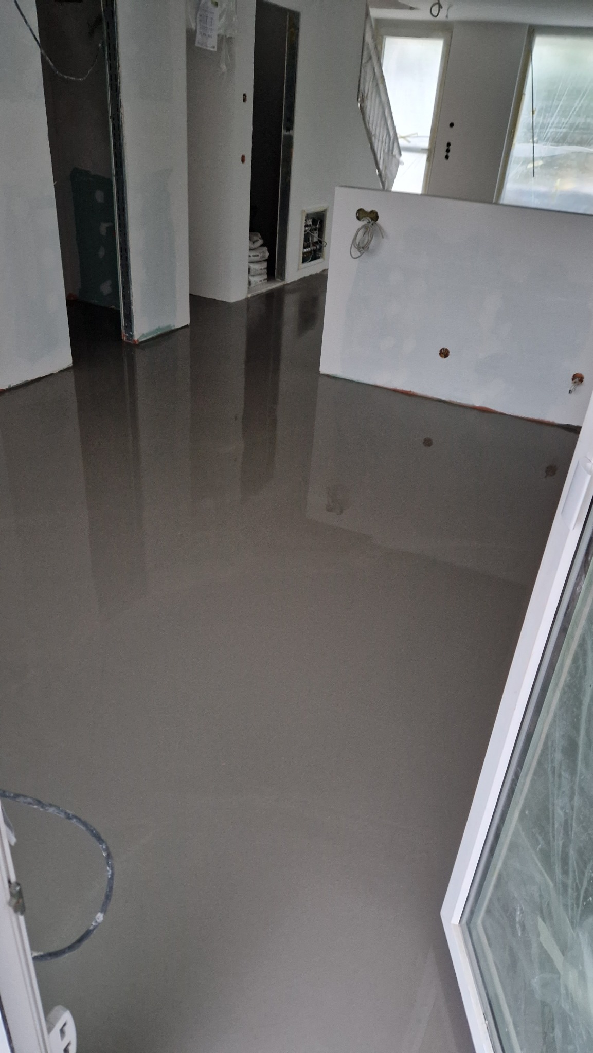 Wylany samopoziomujący podkład podłogowy w jasnym pomieszczeniu w trakcie remontu. Widoczne ściany, otwory drzwiowe i okno. Na podłodze widoczne refleksy światła.