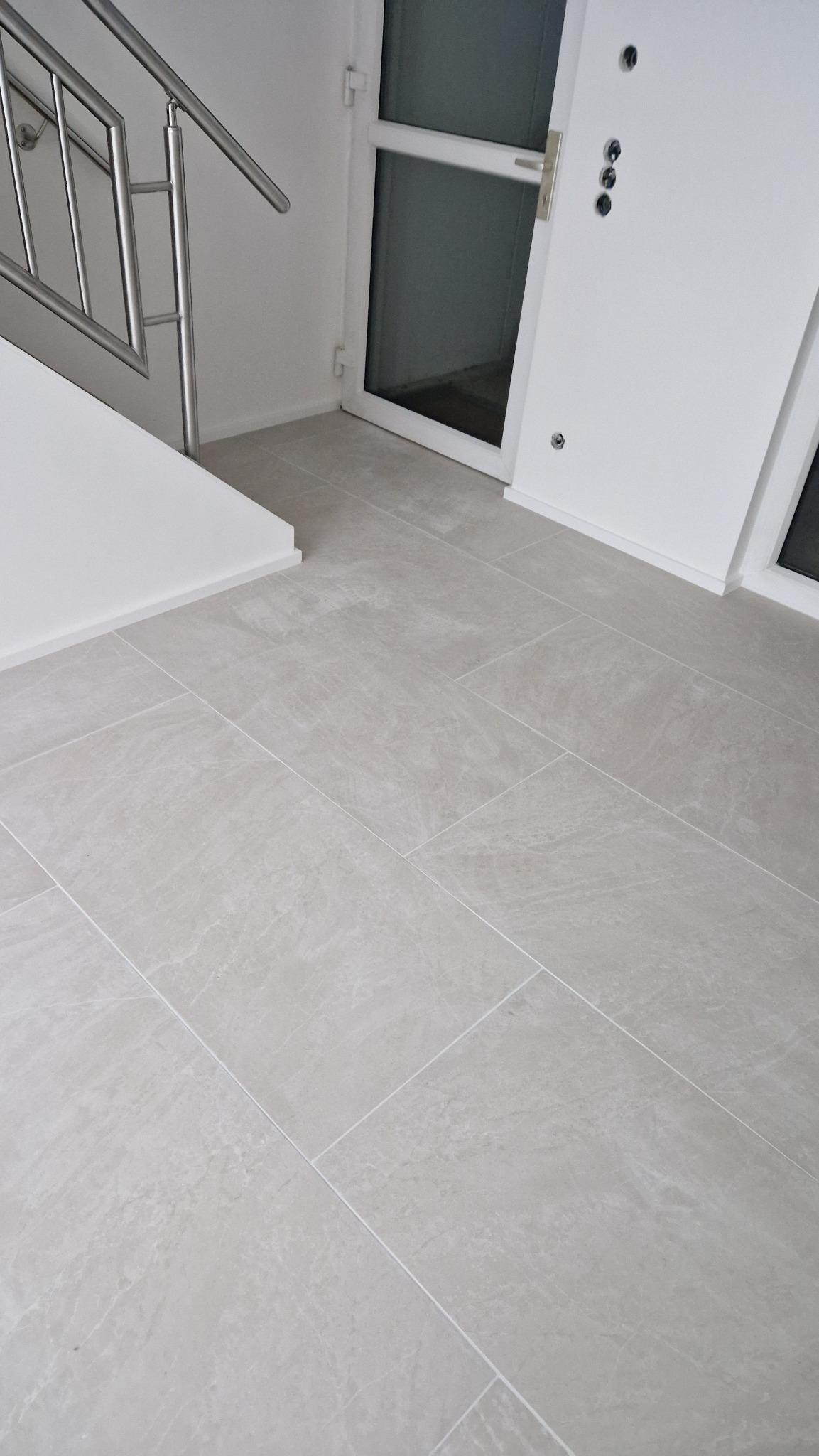 Jasnoszare płytki podłogowe ułożone w korytarzu z metalową balustradą schodów i białymi drzwiami wejściowymi. Minimalistyczny design, czyste linie i neutralna kolorystyka.