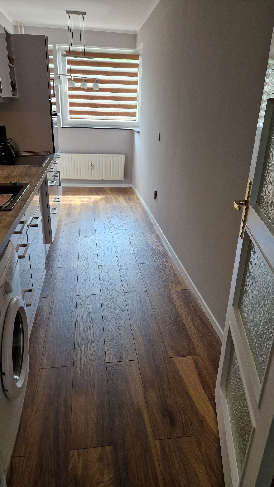 Nowoczesne wnętrze: parkiet imitujący drewno w kuchni, widok z perspektywy podłogi, z fragmentem pralki i otwartych drzwi. Jasne ściany i okno z żaluzjami.