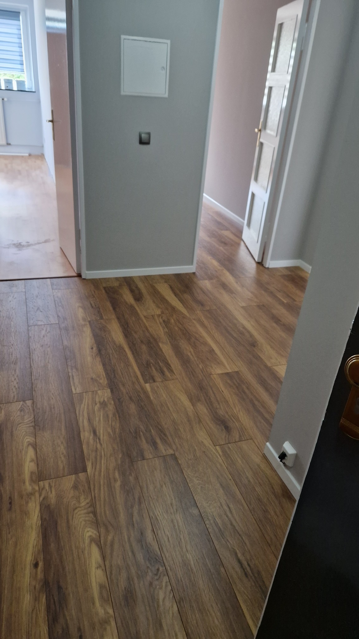 Ukończone układanie paneli podłogowych w odcieniach brązu, widok na korytarz z dwiema parami drzwi, szare ściany z białymi listwami przypodłogowymi i gniazdkiem.