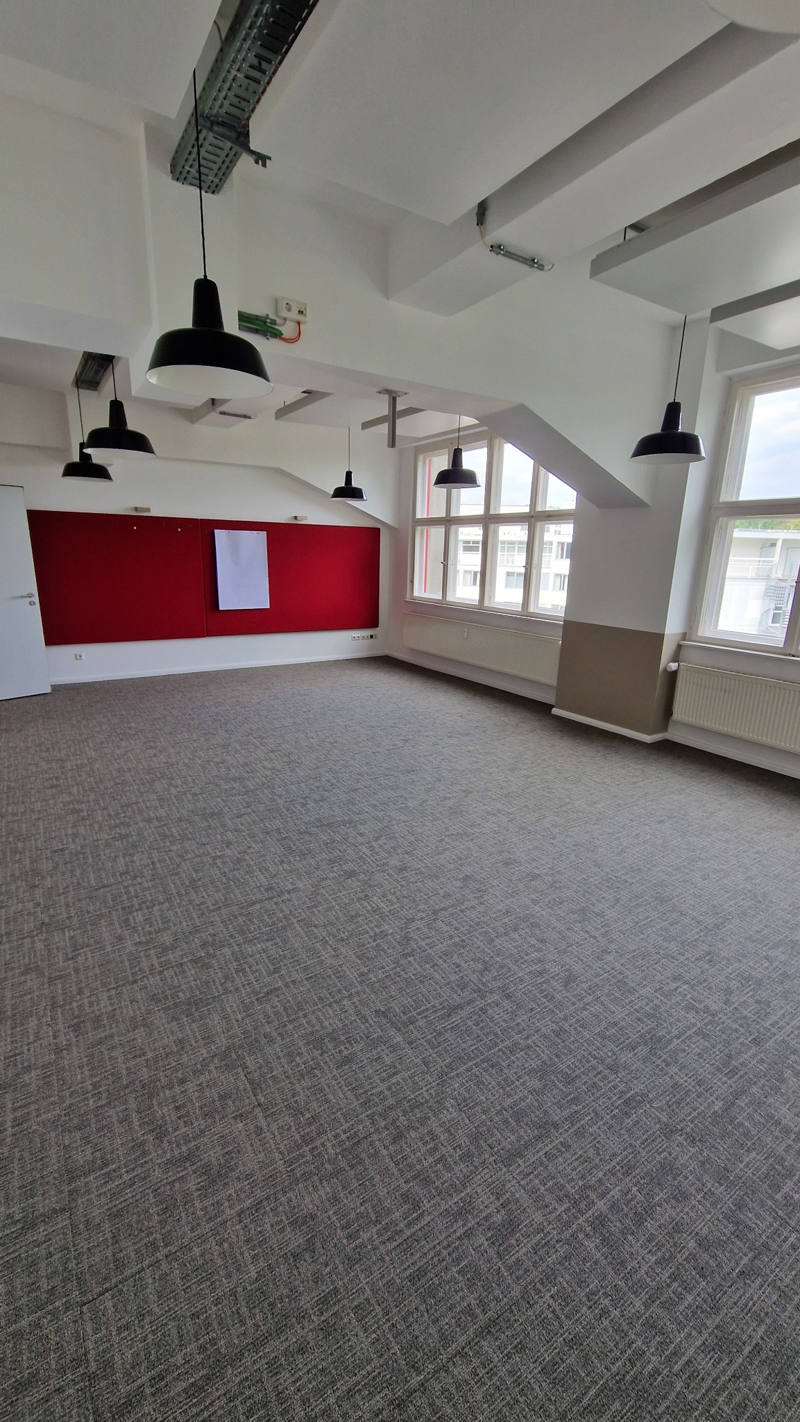 Przestronne biuro z szarą wykładziną dywanową, czerwonym panelem ściennym i wiszącymi lampami. Widok na okna z grzejnikami. Białe ściany i sufit z instalacjami.