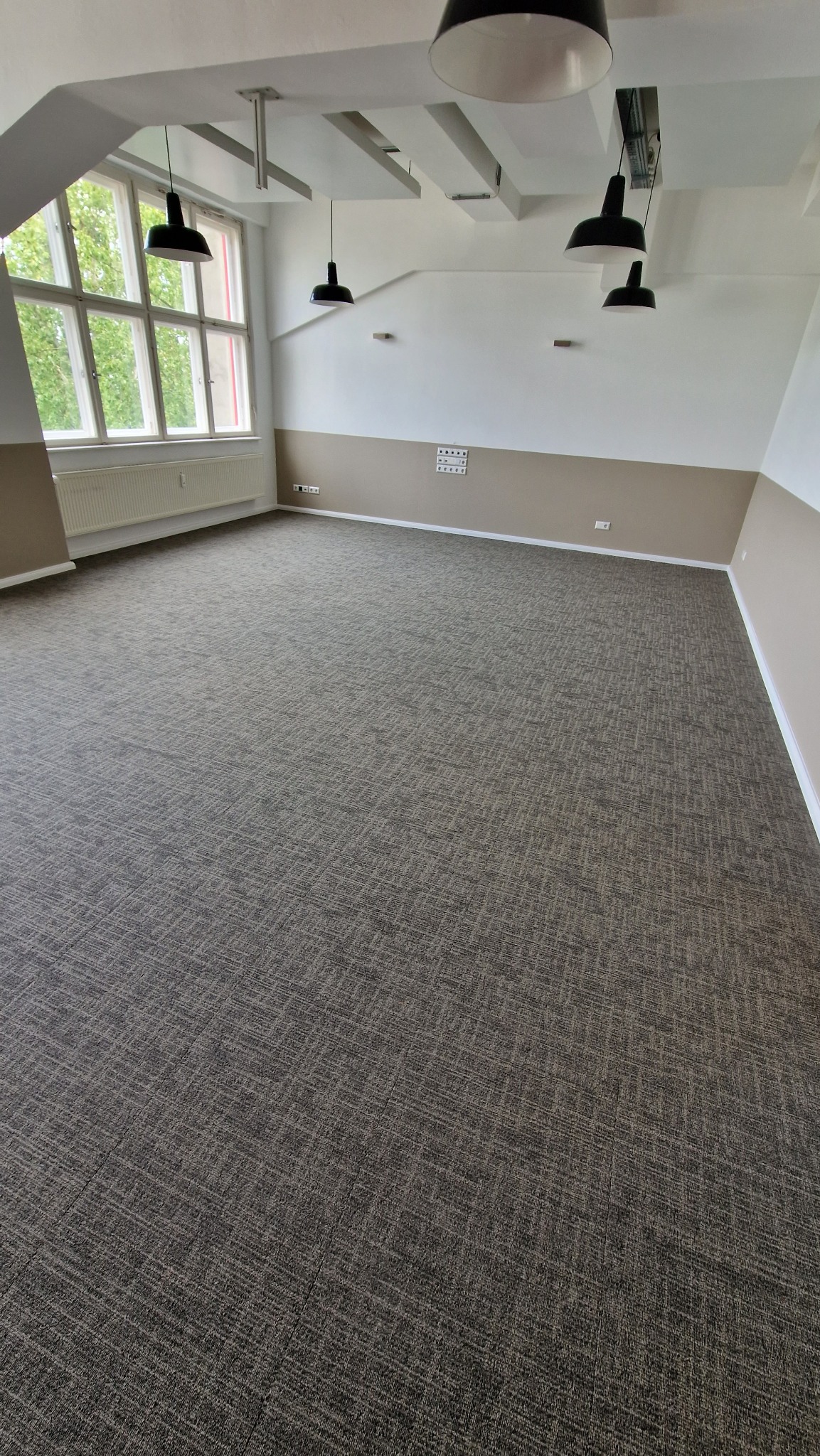 Przestronne wnętrze z wykładziną dywanową w odcieniach szarości, oświetlone wiszącymi lampami. Widoczne okno z widokiem na zieleń i dwukolorowe ściany.