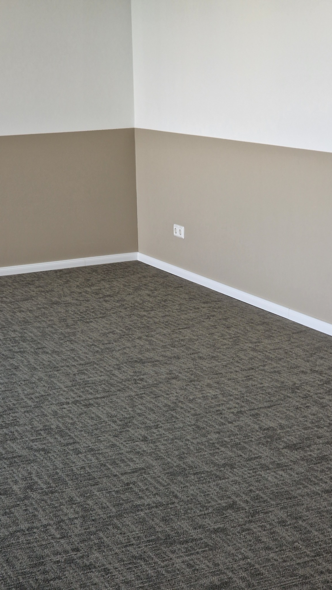 Wnętrze po remoncie: dwukolorowe ściany (beż i biały) z listwami przypodłogowymi i szarą wykładziną dywanową w geometryczny wzór, gniazdko elektryczne na ścianie.