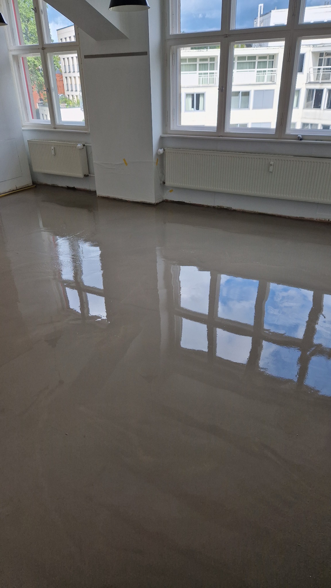 Świeżo wylana, samopoziomująca wylewka podłogowa w jasnym pomieszczeniu z dużymi oknami, odbijającymi błękit nieba. Widoczne grzejniki i przygotowane ściany.