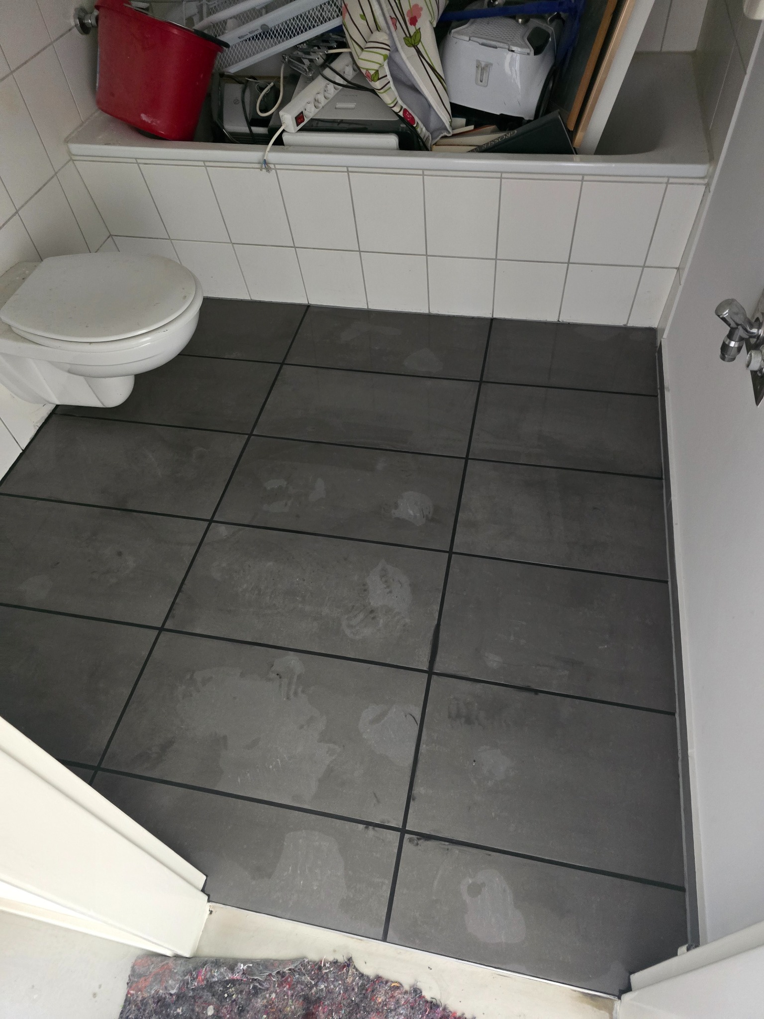 Szare, matowe płytki podłogowe w łazience, widok z góry. Uwagę zwracają ślady na płytkach. Częściowo widoczna toaleta i wanna z akcesoriami.