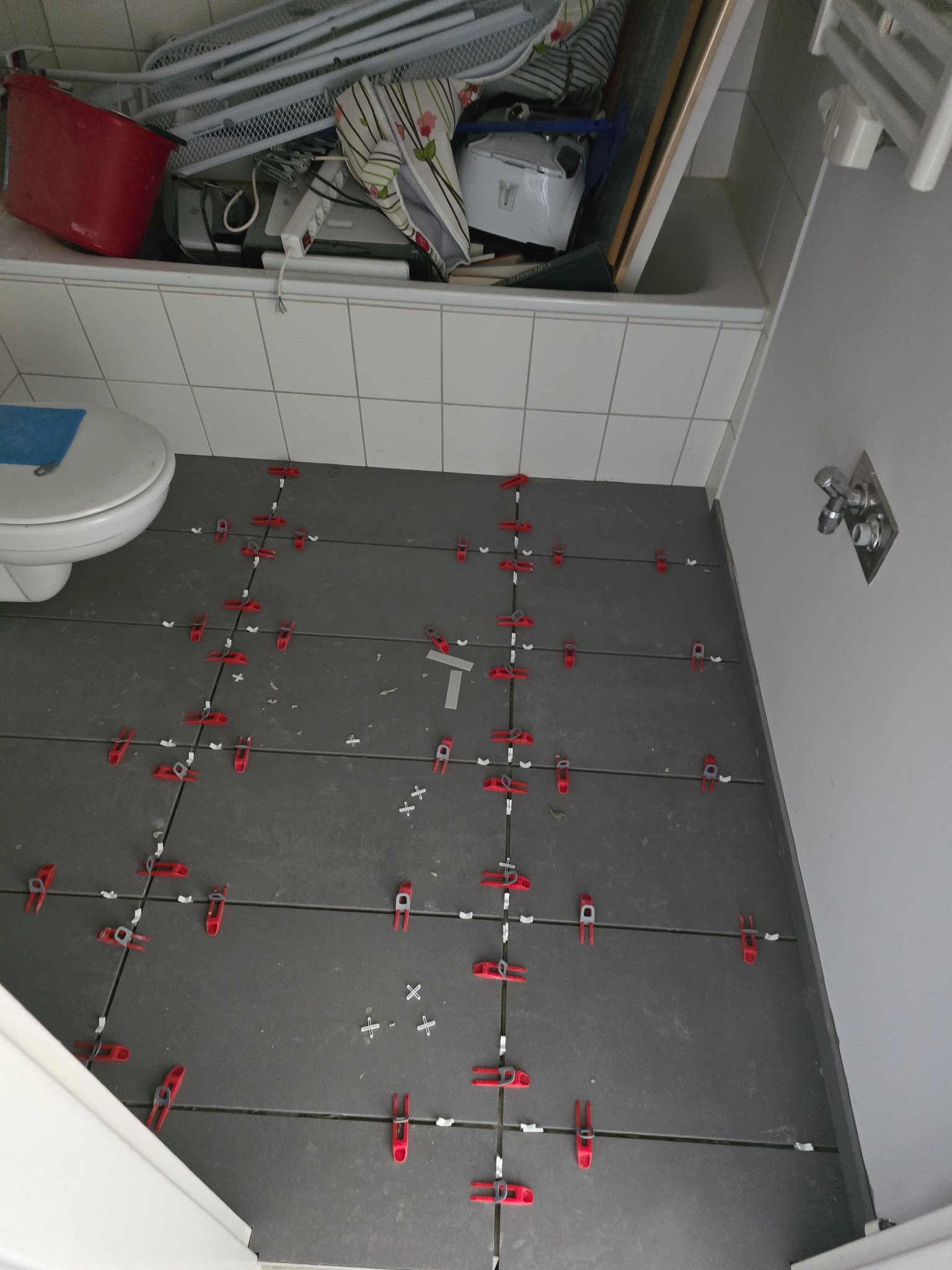 Szare płytki podłogowe w łazience, w trakcie układania, z czerwonymi klipsami poziomującymi, widok z góry, blisko toalety i wanny z akcesoriami.