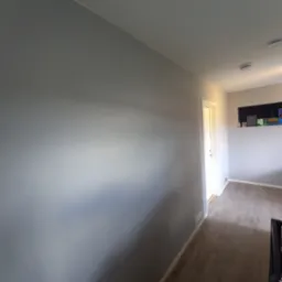 Ściana pomalowana na szaro, widoczna podłoga i drzwi w perspektywie, oświetlenie sufitowe. Minimalistyczny wystrój wnętrza.