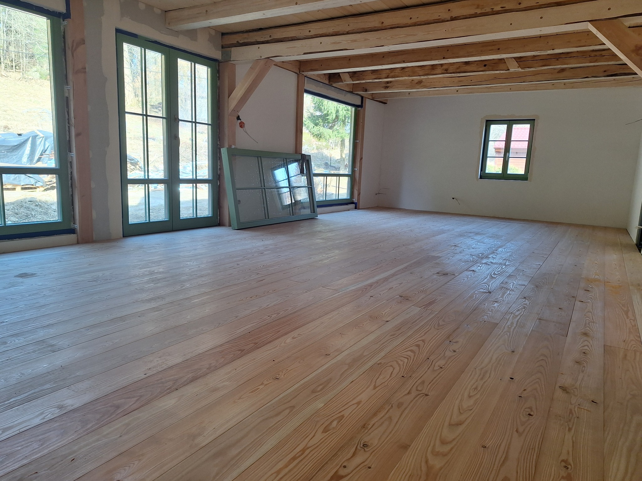 Jasne drewniane deski podłogowe w przestronnym, jasnym pomieszczeniu z dużymi oknami i surowym drewnianym stropem. Okno o zielonej ramie oparte o ścianę.