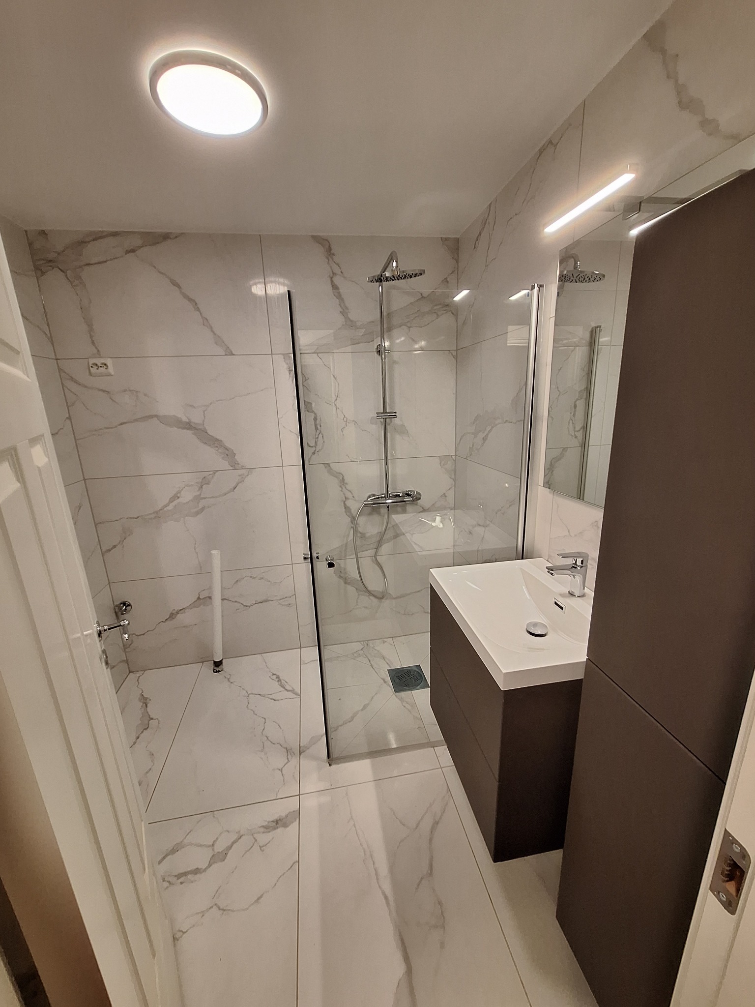 Nowoczesna łazienka z kabiną prysznicową typu walk-in, umywalką w zabudowie i marmurowymi płytkami. Oświetlenie sufitowe i lustro z podświetleniem LED.
