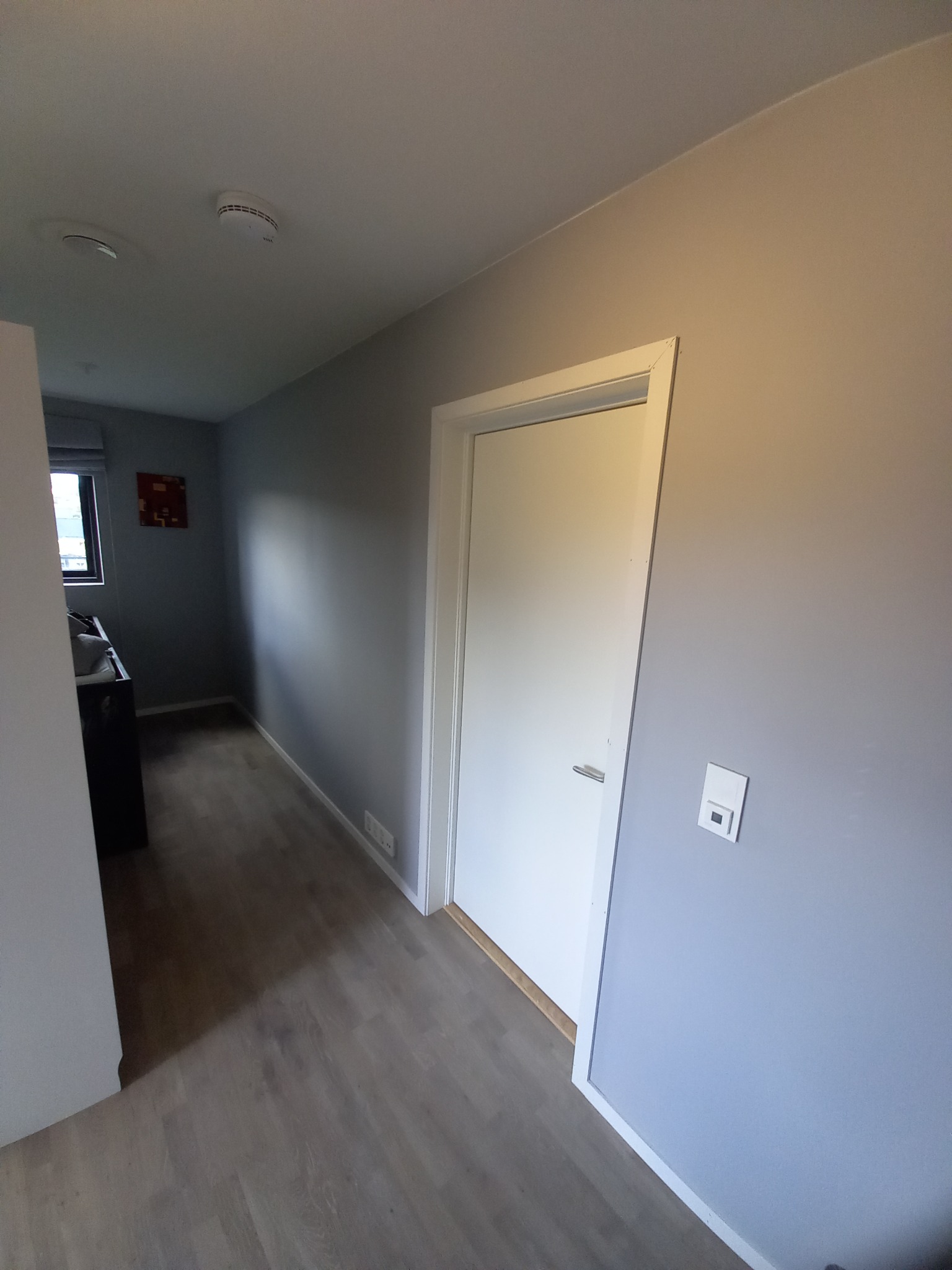 Szary korytarz z białymi drzwiami i drewnianą podłogą. Na ścianie widoczny włącznik światła. W oddali pokój z oknem i obrazem na ścianie.