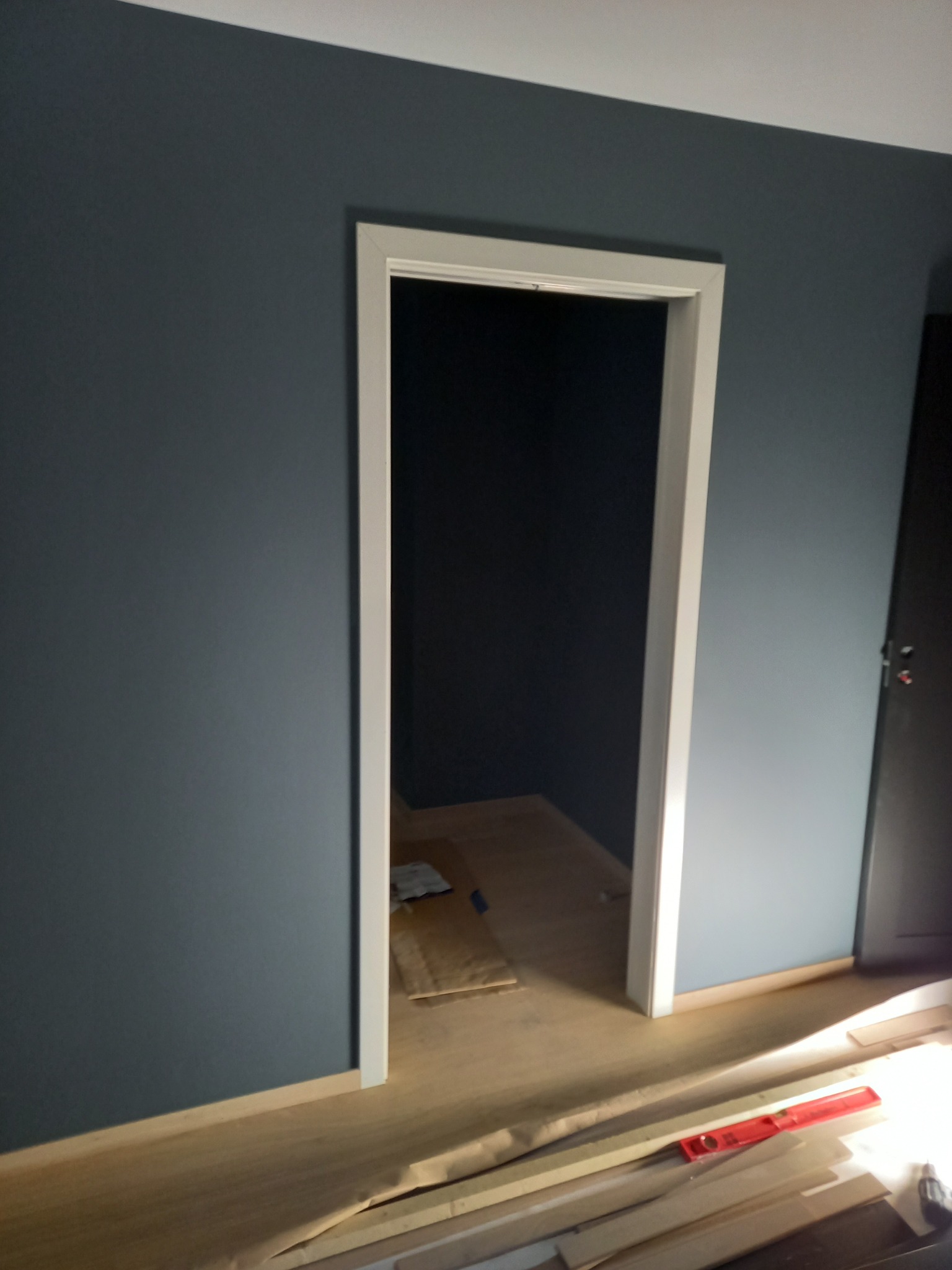 Ściana pomalowana na granatowo z białymi futrynami drzwi. Na podłodze zabezpieczenia i listwy przypodłogowe. Widoczny poziomica.