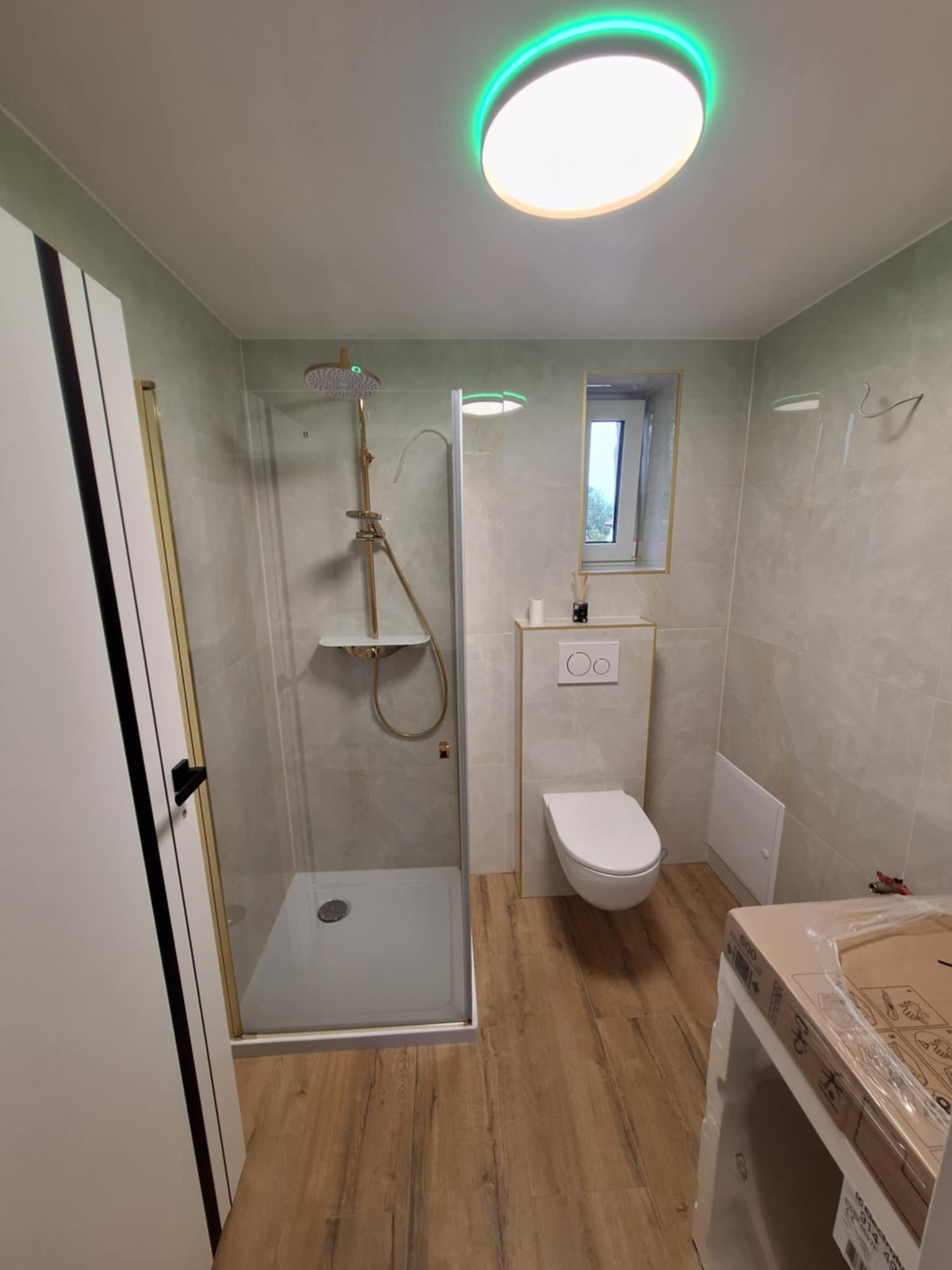 Nowoczesna łazienka po remoncie: kabina prysznicowa ze złotymi akcentami, podwieszana toaleta, drewnopodobne płytki, oświetlenie LED. Widok na okno.