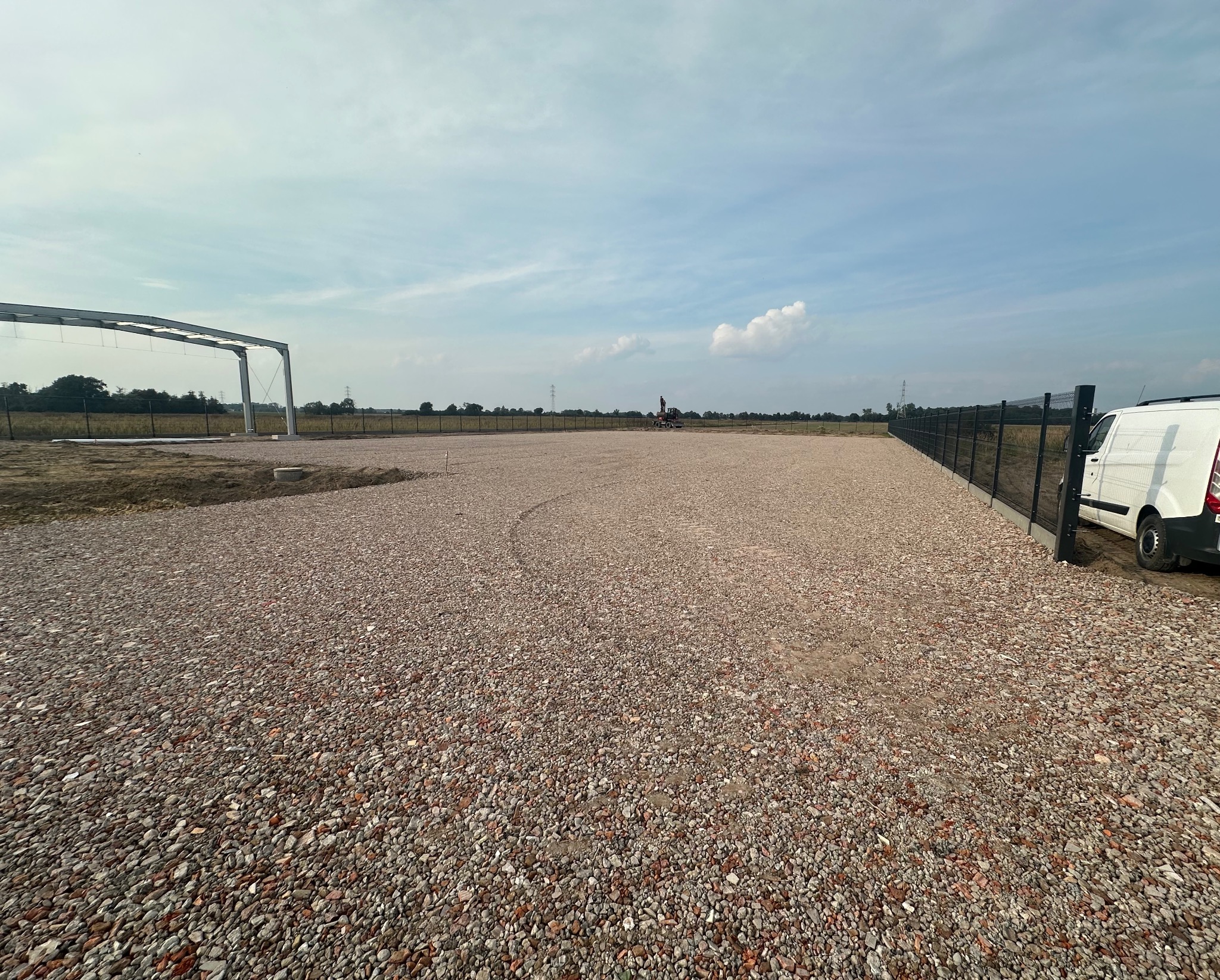 Utwardzony teren żwirowy z widoczną konstrukcją hali w tle, ogrodzeniem panelowym i zaparkowanym busem. Prace ziemne zakończone, widok na rozległy plac.