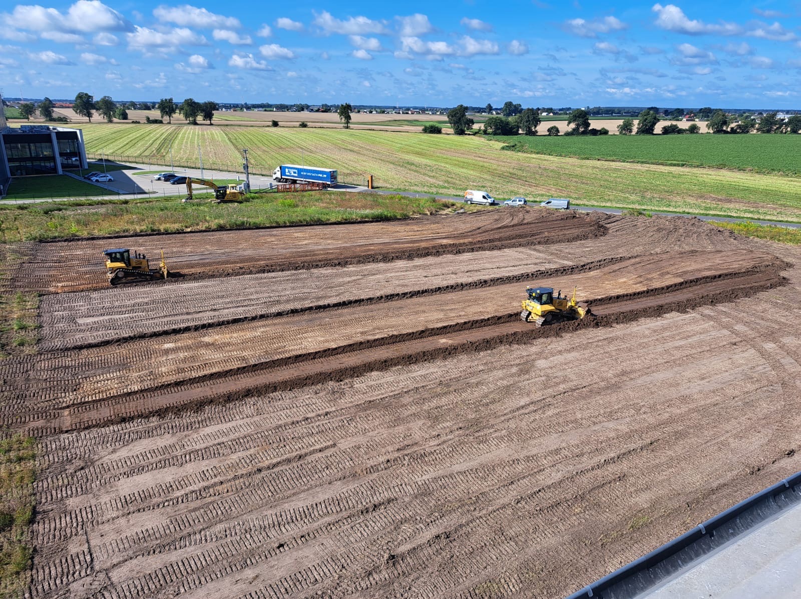 Żółty buldożer w trakcie prac ziemnych na polu, widok z góry. W tle budynek, pola uprawne i błękitne niebo z chmurami. Ślady opon na ziemi.