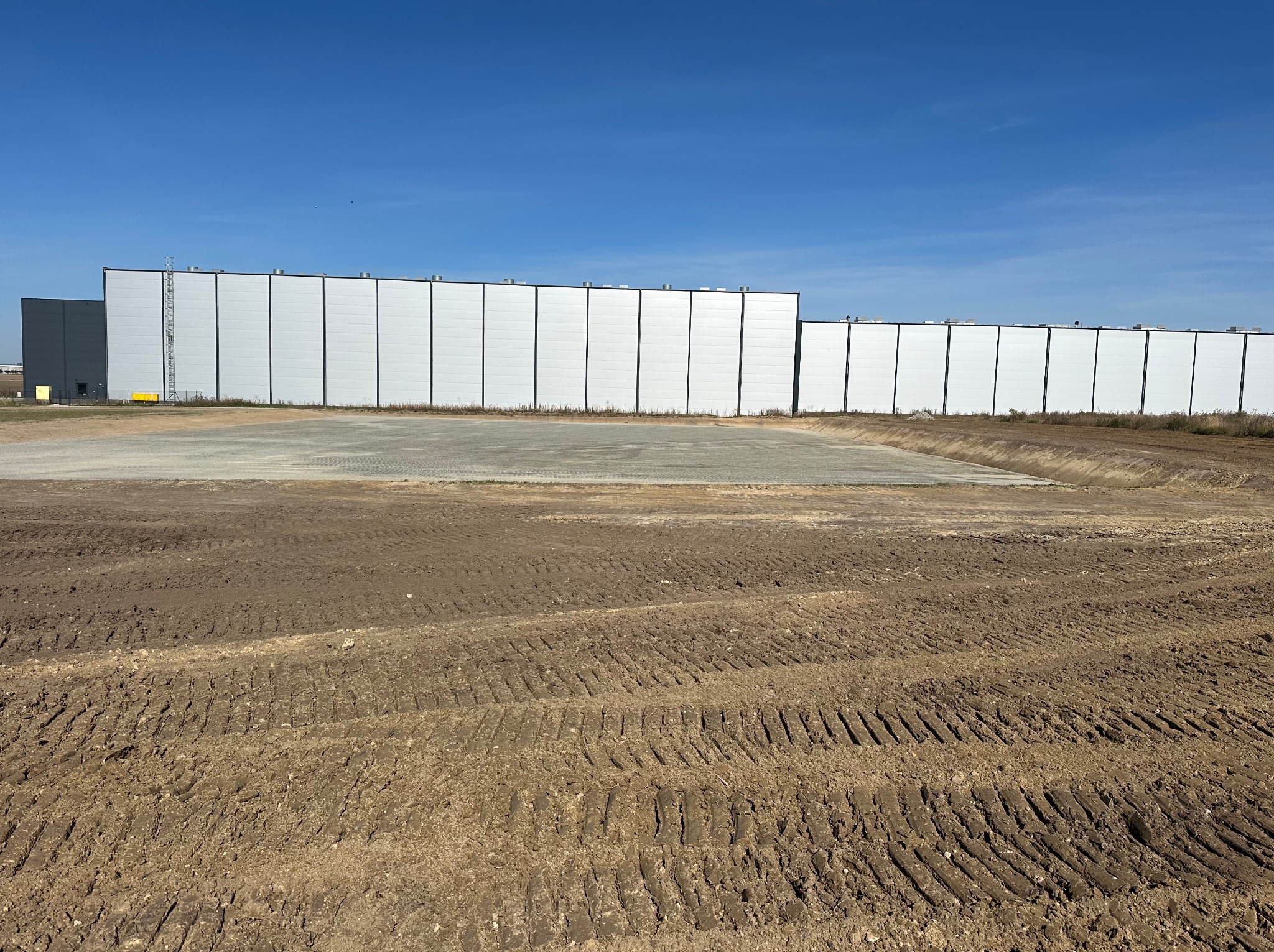 Przygotowany teren pod budowę hali magazynowej, widoczne ślady opon maszyn budowlanych na ziemi, w tle biała hala magazynowa pod błękitnym niebem.