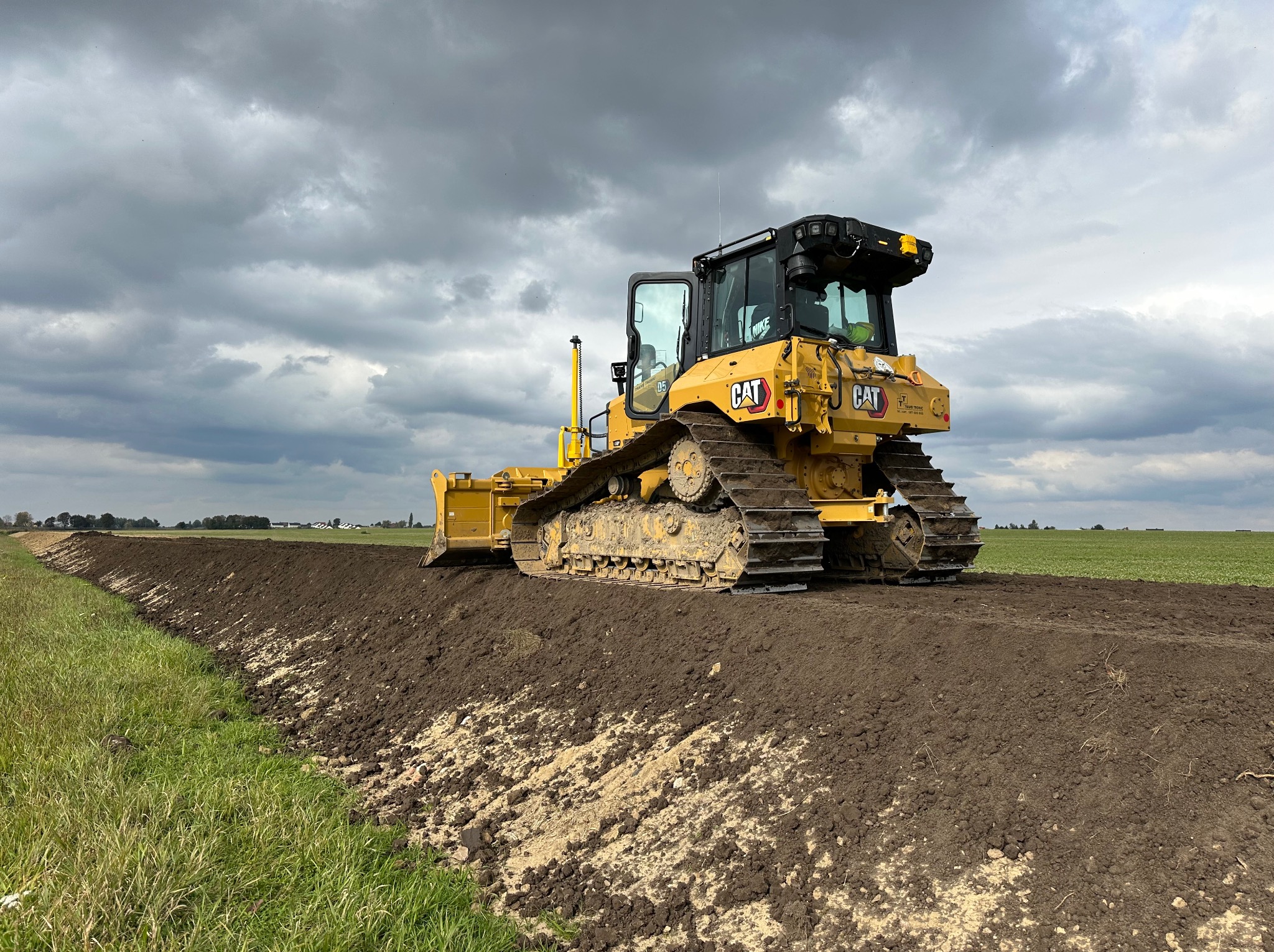 Żółty spychacz CAT na polu, wyrównuje teren pod pochmurnym niebem. Widoczny świeżo wyrównany pas ziemi i zielone pola w tle. Prace ziemne w toku.