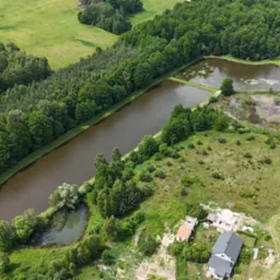 Panoramiczny widok z lotu ptaka na malownicze stawy rybne otoczone lasami i zabudowaniami, w słoneczny dzień. Widoczna gra światła i cienia na wodzie i zieleni.