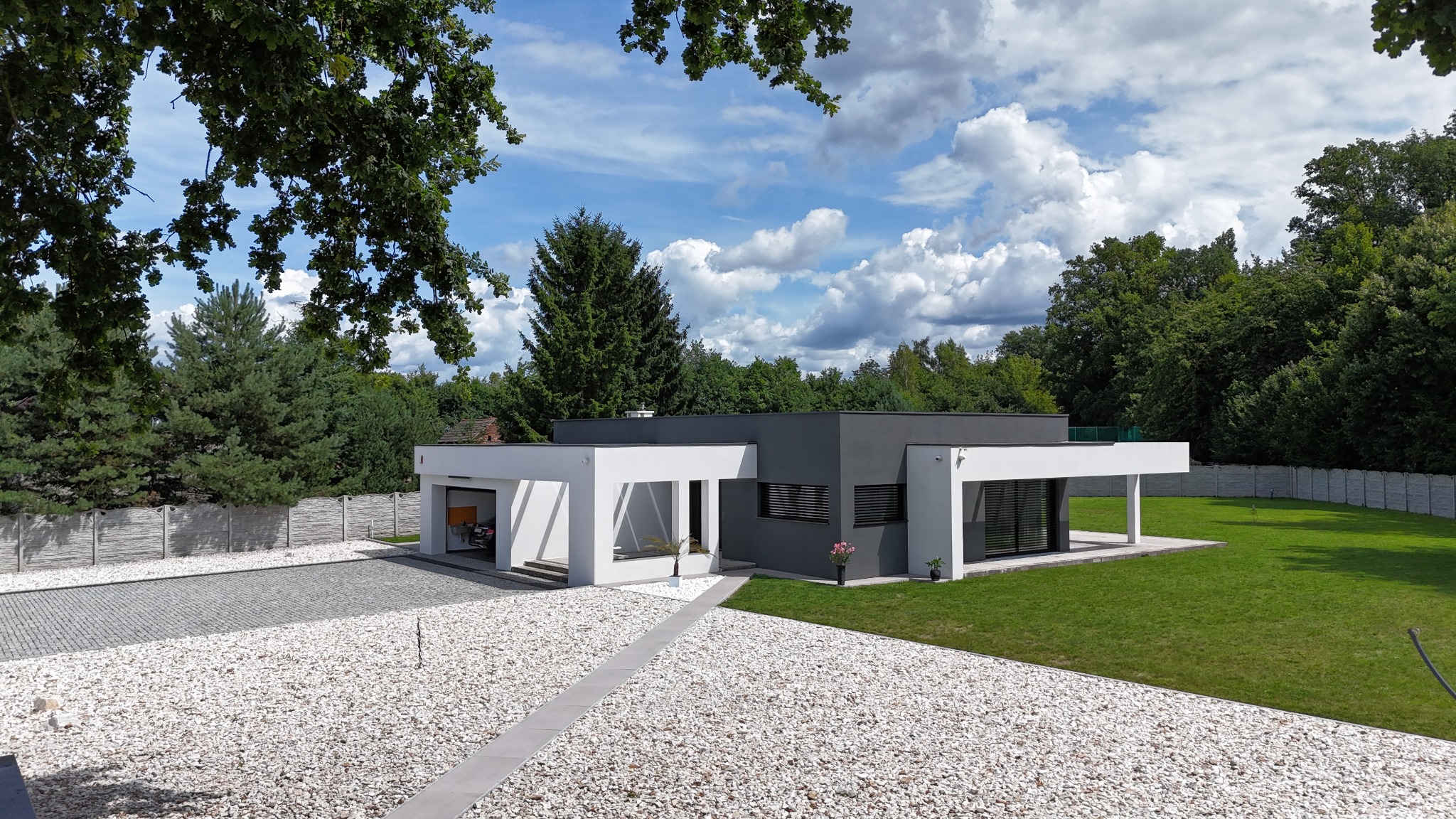 Nowoczesny dom jednorodzinny z płaskim dachem, szarą elewacją i białymi elementami architektonicznymi, otoczony zielenią i żwirowym podjazdem w słoneczny dzień.