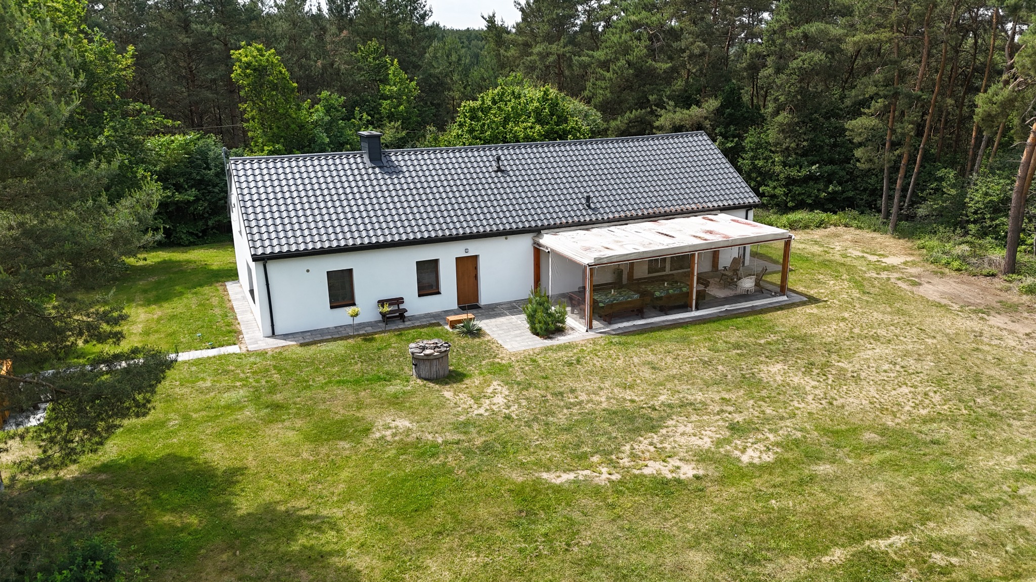 Biały dom z szarą dachówką, widziany z góry, z zadaszonym tarasem i studnią na trawniku, otoczony drzewami. Ujęcie podkreśla architekturę i zagospodarowanie terenu.