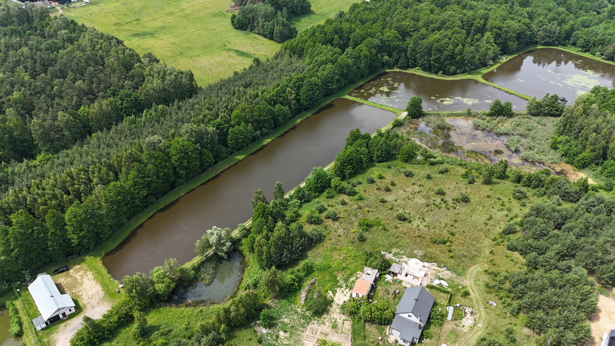 Panoramiczny widok z lotu ptaka na malownicze stawy rybne otoczone lasami i zabudowaniami, w słoneczny dzień. Widoczna gra światła i cienia na wodzie i zieleni.
