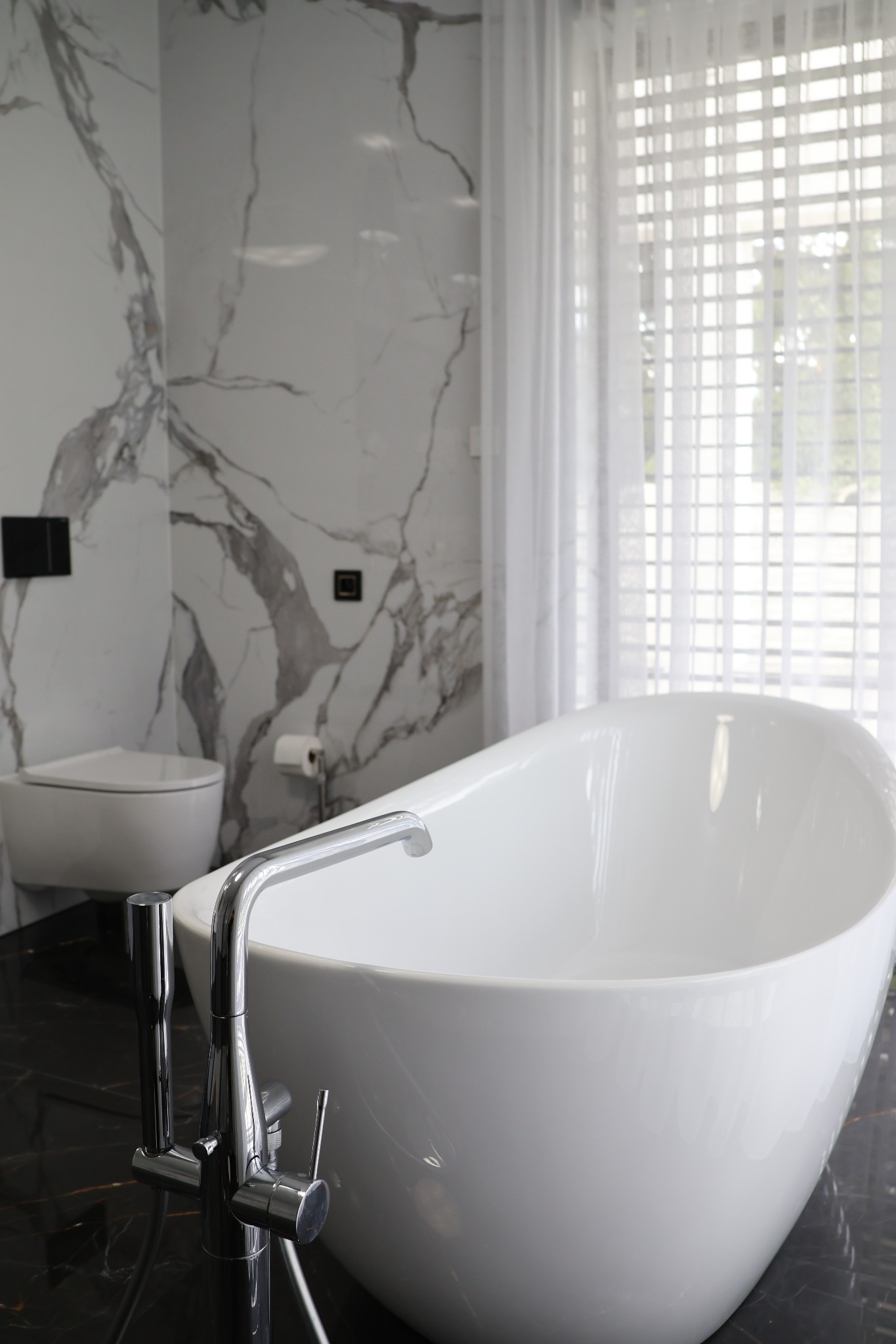 Elegancka, wolnostojąca wanna w nowoczesnej łazience z marmurowymi ścianami i delikatnymi, białymi zasłonami w oknie. Bateria wannowa chromowana.