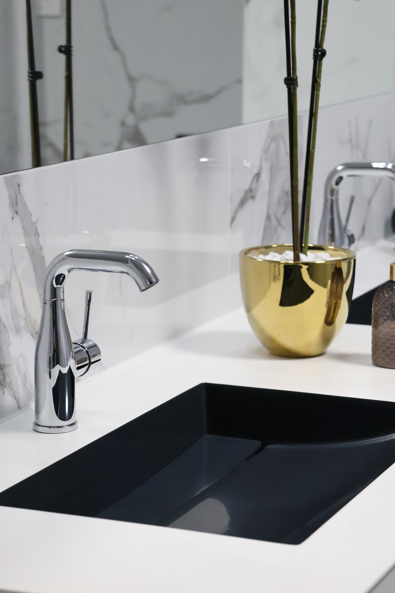 Nowoczesna łazienka: chromowana bateria umywalkowa, czarna, prostokątna umywalka, biały blat z marmurowym tłem i złota doniczka z roślinami.